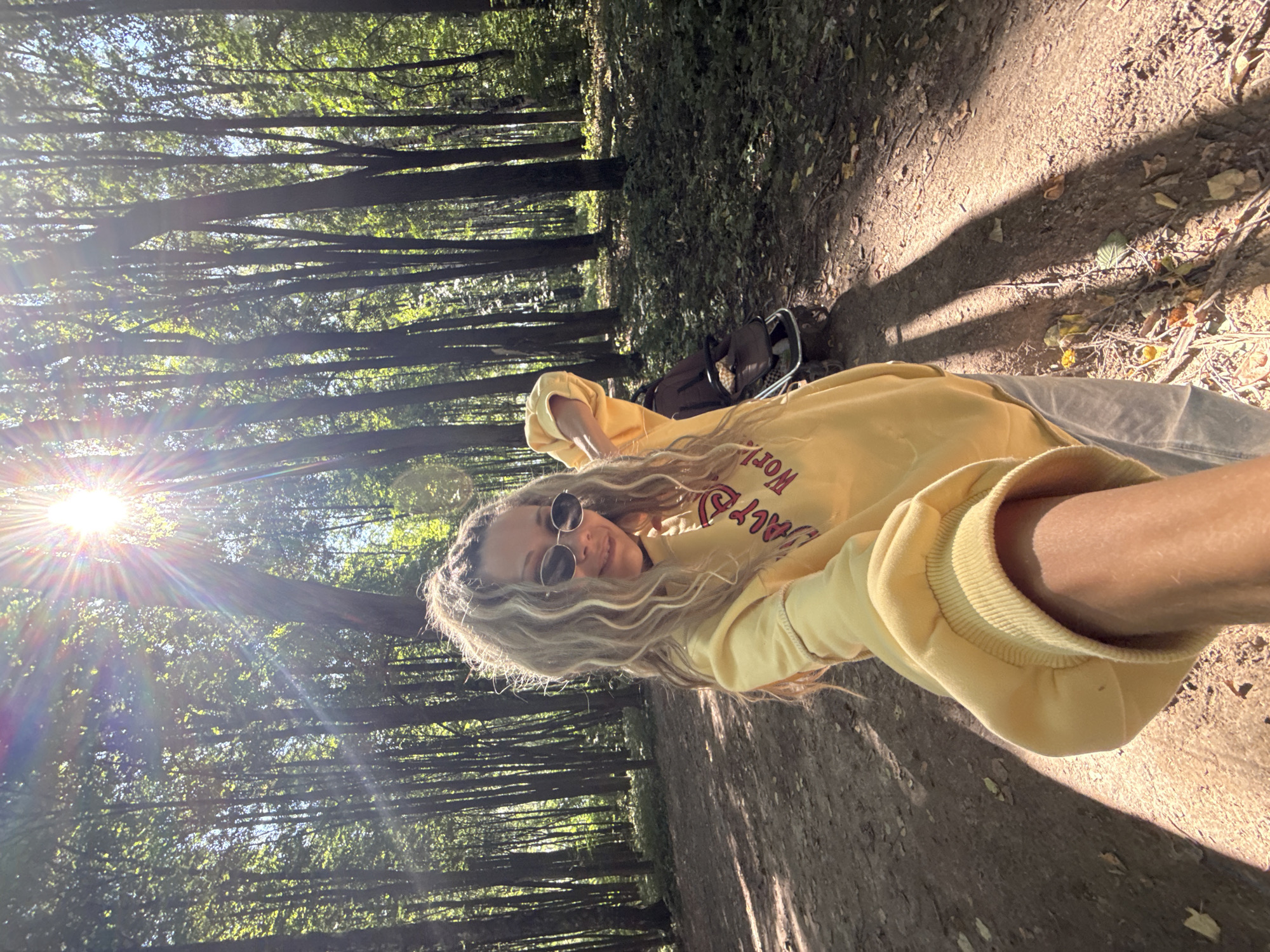 An outdoor, waist-up, selfie-style medium shot captures a woman with long, wavy blonde hair, pulled into a ponytail, wearing round sunglasses and a yellow hooded sweatshirt with red lettering. She stands on a dirt path in a forest, with tall trees and dappled sunlight visible in the background. Behind her, to the right, is a dark-colored stroller. The sun is prominently featured in the upper left quadrant of the image, with visible lens flare. The woman is smiling and looking towards the camera, with her left arm extended as if taking the photo. The setting appears to be a park or wooded area within or near the city of Rusanovo, Russia, given the presence of mature trees and a well-maintained path.
