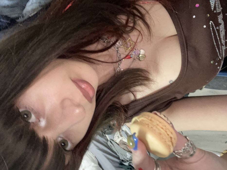 A woman with long, dark hair and light eyes is holding a yellow macaron. She is wearing a brown tank top with embellishments. A collection of necklaces, including one with a Star of David, adorn her neck. Her hand features ornate rings and colorful nail polish. The background appears to be a casual indoor setting, possibly a bed. The photo is taken at a slightly tilted angle.