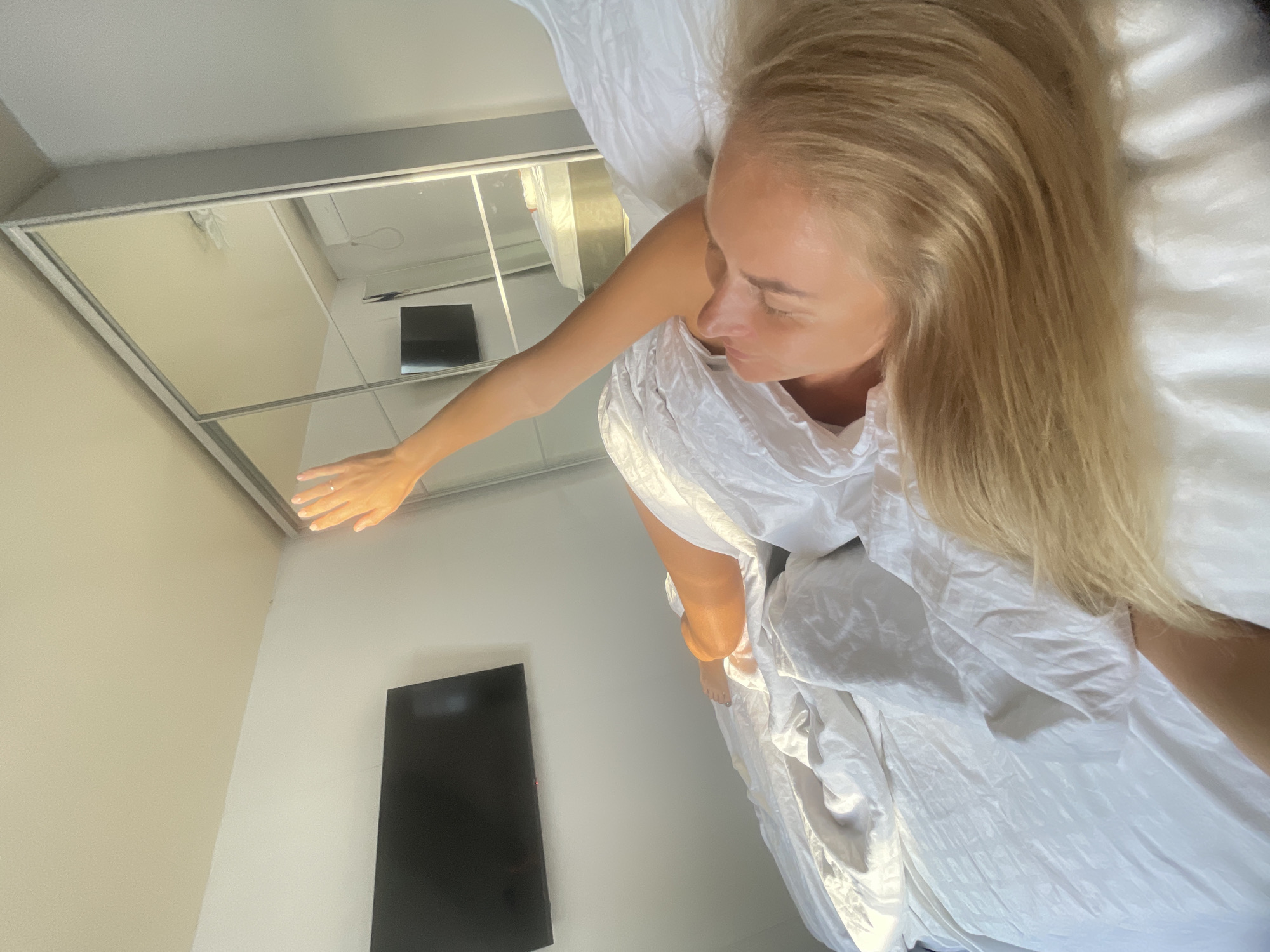 A blonde woman lies on a bed covered in white linens, her body partially obscured by a white sheet. Sunlight streams in from the right, illuminating her face and raised right arm, which is extended towards the light. A ring is visible on her ring finger. In the background, a large mirrored wardrobe reflects the room, including a television mounted on the wall. The overall setting appears to be a bedroom. The location in Baranovka, Russia, is not visually ascertainable from the image.