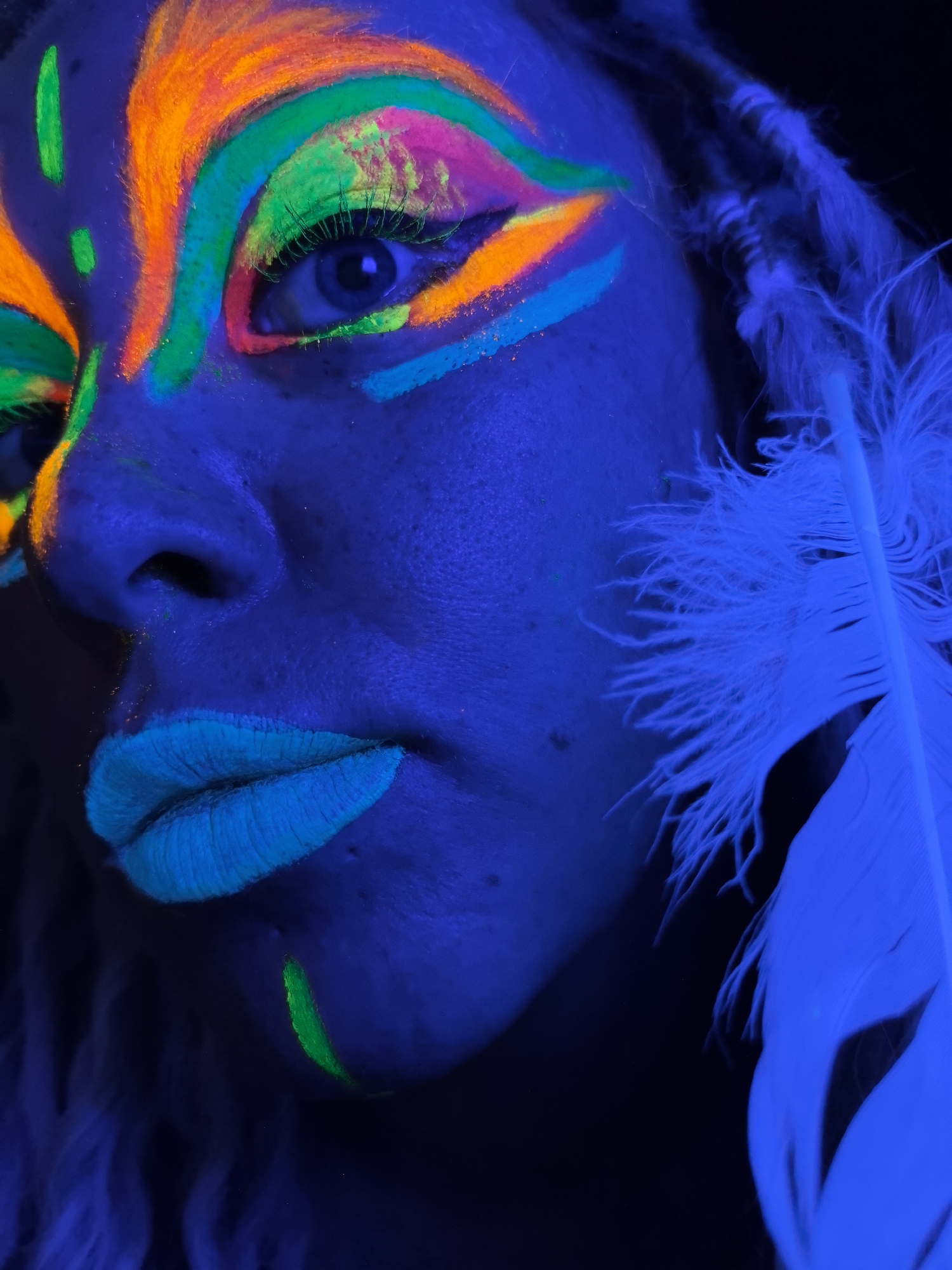 A close-up image reveals a person with fluorescent body paint under a blacklight. The person has orange, green, and pink geometric designs around the eye, and a single orange stripe under the eye. There are green stripes on the forehead and chin. The lips are painted a light blue. A white feather is visible near the right side of the face. The skin appears blue under the light. The background is dark and indistinct. There is no evidence that supports this image being taken in the city of Noviy, Russia.