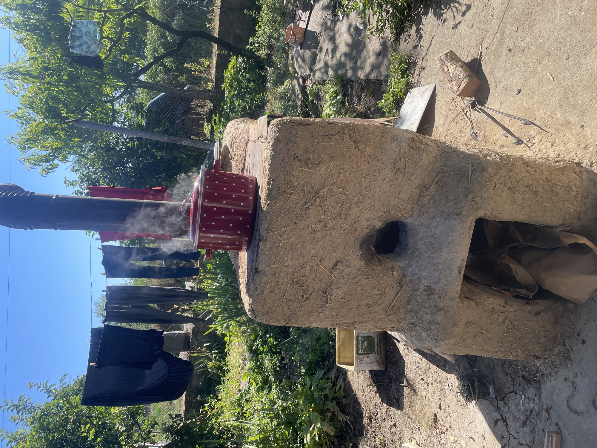 In Zheglartsi, Bulgaria, a rustic outdoor cooking setup is depicted. A large, earth and straw oven stands prominently in the foreground, with a metal pipe extending upwards. A red pot with white polka dots rests on the top of the oven, emitting steam, indicating that food is being cooked. The oven has a lower opening, suggesting it is wood-fired, with some paper or fabric visible inside. To the left of the oven, a white plastic container sits on the ground. Clothes are hung on a clothesline in the background, drying in the sun under a clear blue sky, adding to the rural atmosphere. The area is surrounded by greenery, with trees and bushes, and a fence can be seen in the distance. The ground is a mix of dirt and concrete, with scattered twigs and a pair of metal tongs lying near the oven. The scene suggests a traditional way of life, possibly in a village setting.