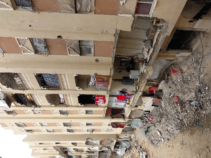 This image depicts a street scene in Khan Yunis, Palestinian Territory, characterized by extensive structural damage to multiple multi-story residential buildings. The buildings are constructed primarily from beige and brown stucco over concrete, featuring ornate white balcony railings and window surrounds, many of which are now damaged or partially missing.
The foreground and ground level between the buildings are covered in a dense layer of rubble, consisting of broken concrete, rebar, dirt, and miscellaneous debris. Portions of the buildings, particularly on the lower and mid-levels, have sustained severe damage, with entire sections of walls collapsed, exposing interiors. Reinforced concrete slabs are visible where floors have been compromised.
Evidence of continued habitation exists amidst the destruction. Laundry, including red, white, and patterned fabrics, hangs on lines and over railings on multiple balconies and exposed interior sections across different floors. On one exposed mid-level floor, a black water tank and a white washing machine are visible. On an upper-level balcony behind a decorative metal grille, a person wearing a yellow garment is partially discernible.
The damage to the building facades includes numerous holes and impacts, broken or boarded-up windows, and charred areas around some openings. The structural integrity appears compromised in many sections, with rebar exposed. The sky above is overcast or bright and indistinct. No active events or direct interactions between individuals are visible in the static scene.