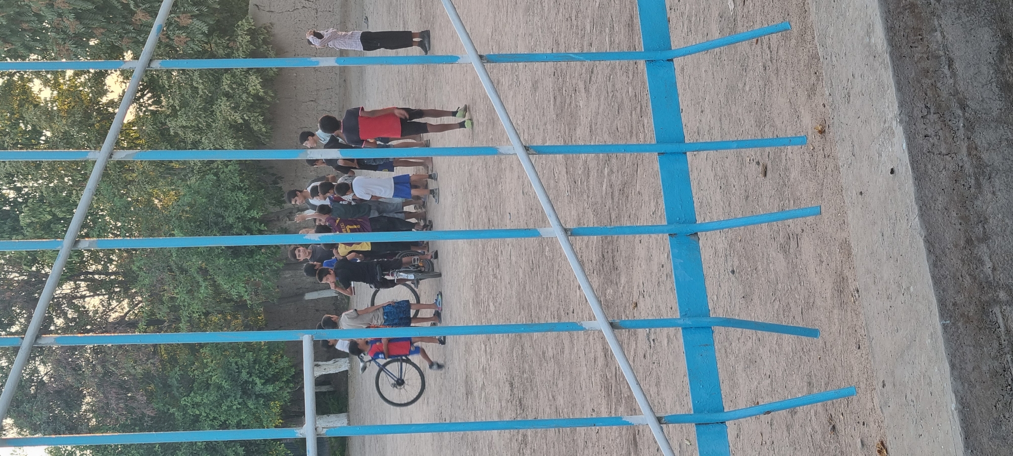 A group of approximately 15 boys and young men are gathered on a dirt field in a residential area of Хаваскор, Tajikistan. The scene is viewed through a blue metal fence. Some of the individuals are wearing athletic clothing. Two bicycles are present on the field, one large and one small. The group appears to be interacting casually, with some standing and talking while others are observing. The environment includes trees and a wall in the background, suggesting an outdoor, possibly recreational, setting. The lighting indicates it is daytime, likely late afternoon or early evening, given the soft sunlight.