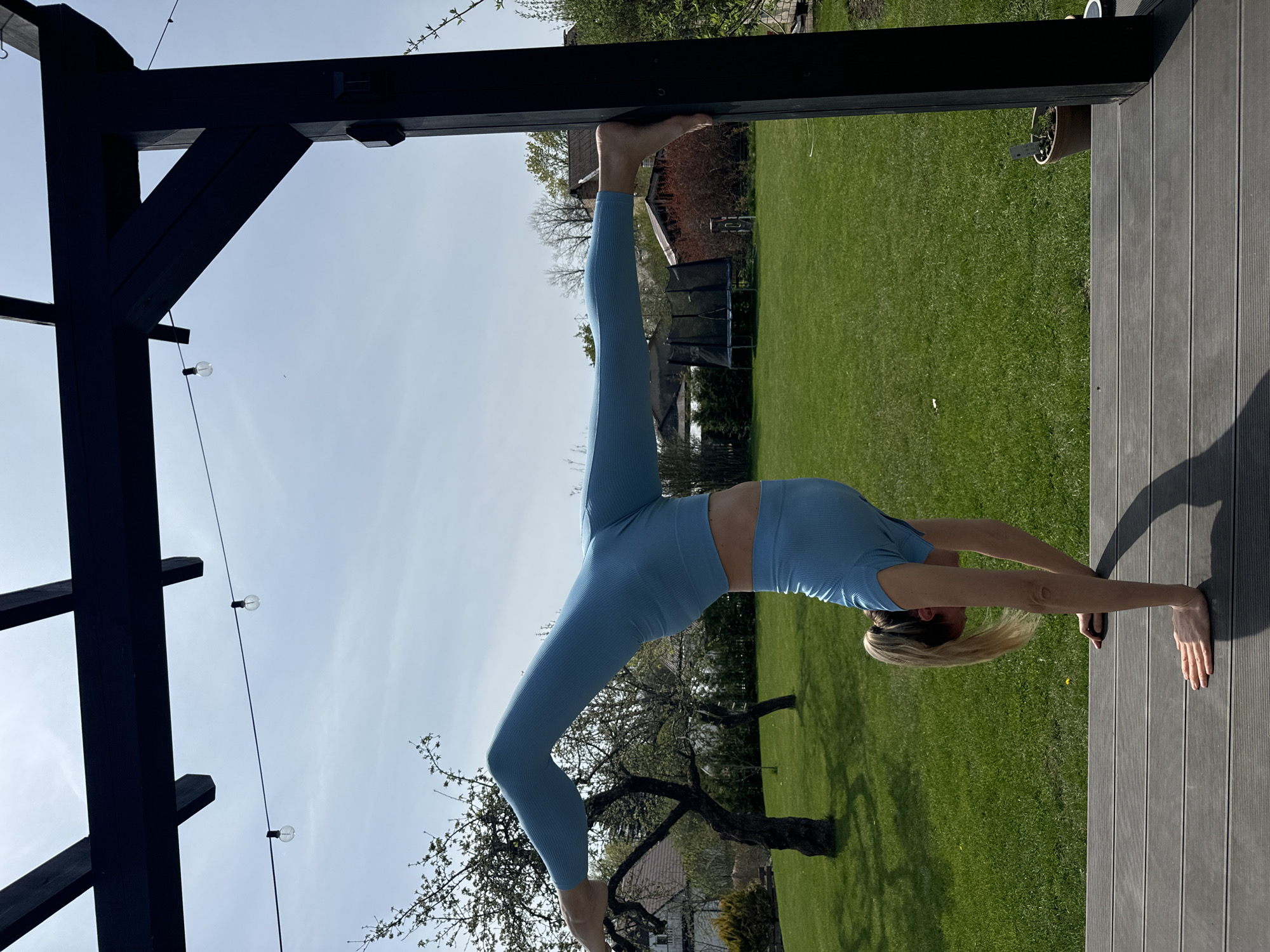 The image depicts a person, presumably female, performing a yoga pose outdoors. She is positioned upside down, with one foot gripping a dark, wooden beam structure and her hands placed on a grey, wooden deck. Her body forms an arch, with one leg extended vertically upwards. She is wearing light blue athletic attire.
The backdrop consists of a well-maintained green lawn, scattered trees, and glimpses of residential buildings, suggesting a suburban or residential area. Hanging lights are visible along the dark, wooden beam structure. The scene appears to be shot during daylight hours under a slightly overcast sky.
Given the prompt's reference to Carnikava, Latvia, it can be inferred that the location is likely within a residential area of this city.