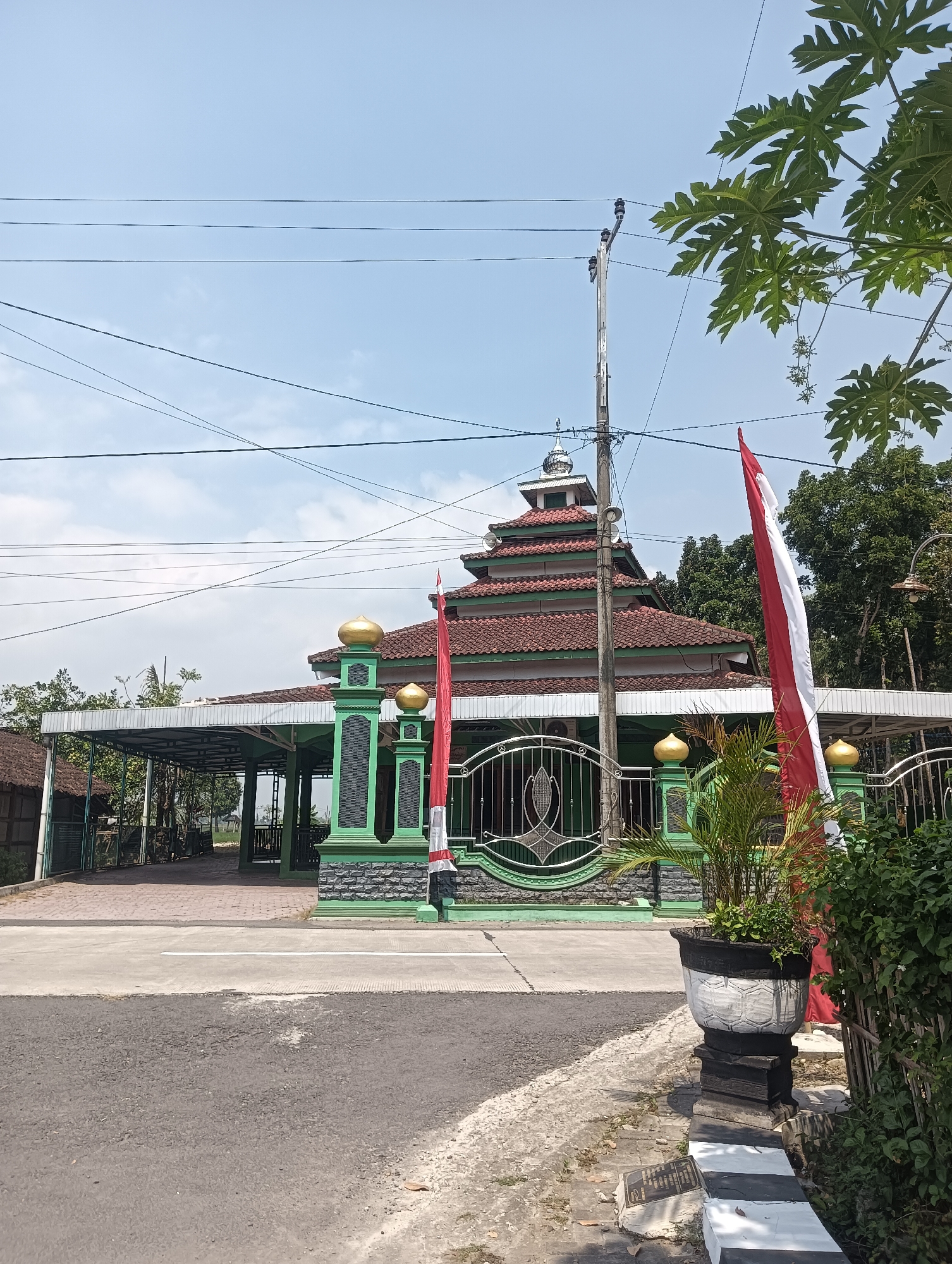 The image shows a mosque with a green facade and a red tiled roof, located on the side of a road in the city of Tanggan, Indonesia. The mosque has several golden domes and is surrounded by a fence with intricate metalwork. In front of the mosque, there are two large Indonesian flags, red and white, planted on flagpoles. A power pole with several electrical wires runs diagonally across the scene, from the upper right to the lower left. A potted plant is visible in the foreground to the right of the road, and a large tree with green leaves is partially visible in the upper right corner. The sky is clear blue with a few scattered clouds. The overall scene depicts a peaceful, sunny day in a rural or semi-rural setting in Indonesia.
