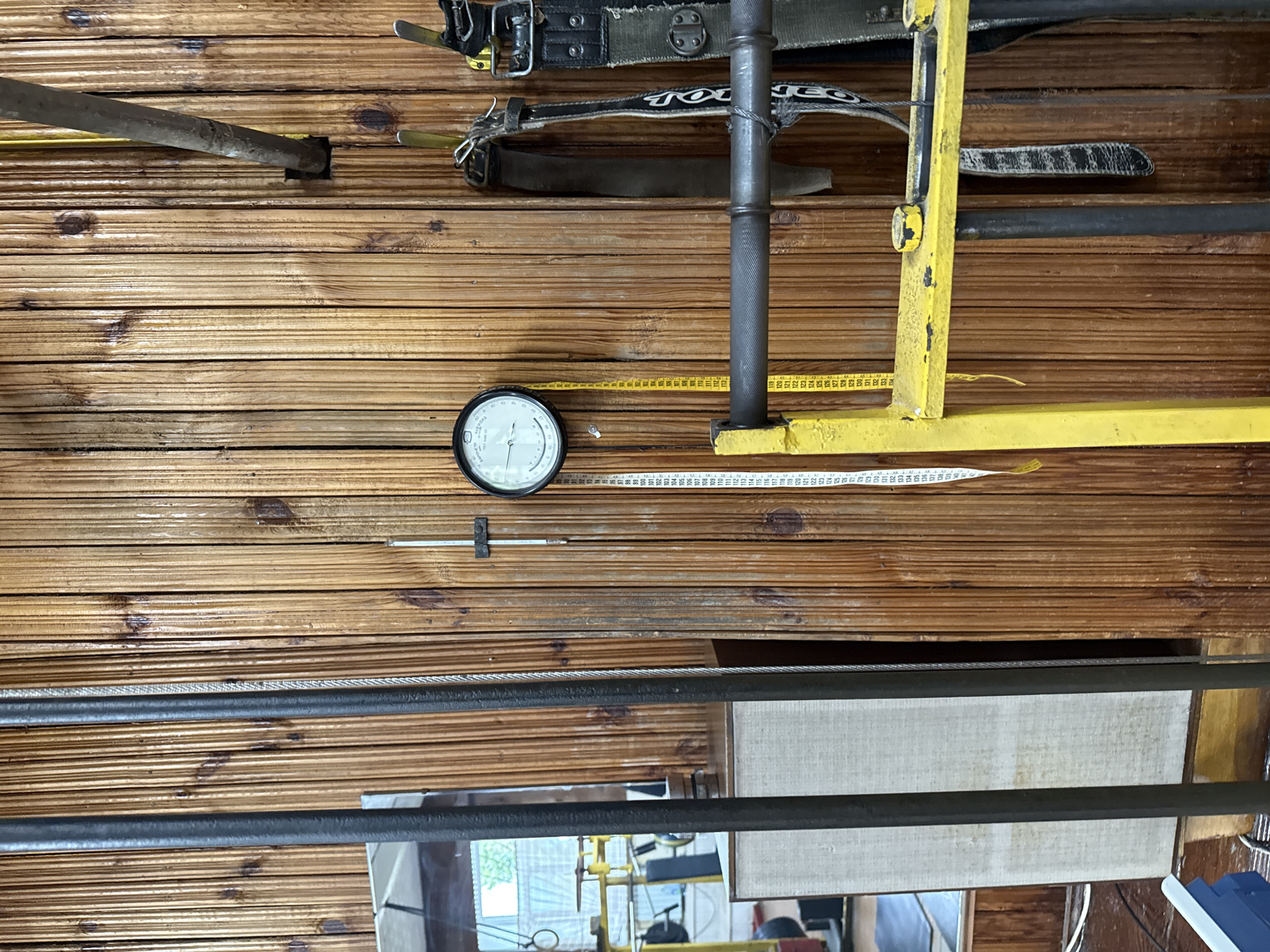 The image is a close-up shot of a gym wall made of vertical wooden planks. Several objects are visible, including a circular gauge with a needle, a yellow measuring tape hanging vertically, and two weightlifting belts. A metal bar, likely part of a weight machine or rack, is also prominent in the foreground. The overall impression is of a utilitarian, potentially older gym environment. The location in the city of Kazan, Russia, is not discernible from the visual elements present in the image. There are no people visible in the frame, and no specific actions or interactions are depicted.