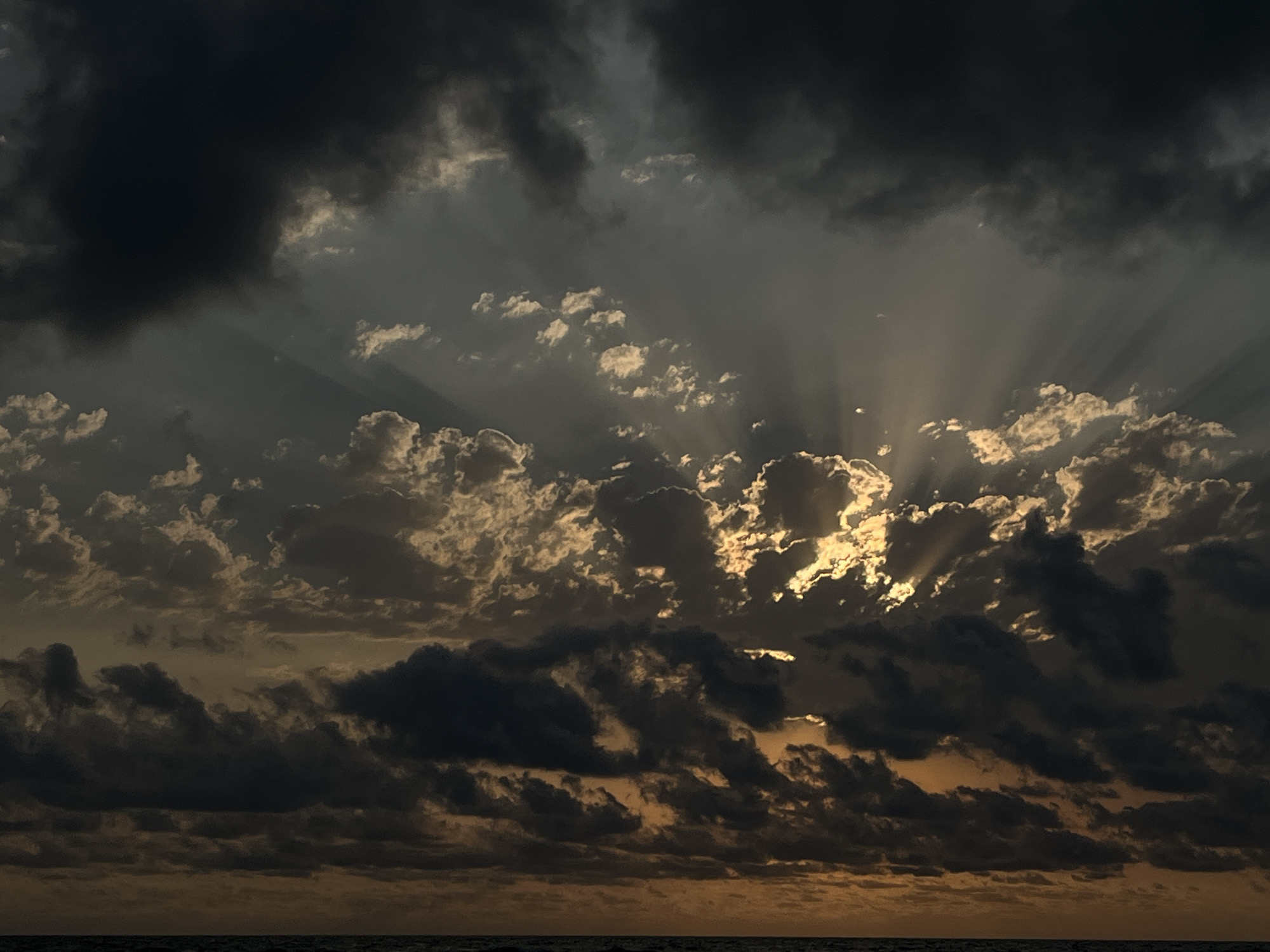 This is a photograph of a dramatic sky over a body of water, likely the Black Sea, near the village of Supsa in Georgia. Dark, heavy clouds dominate the upper portion of the image, with shafts of golden sunlight breaking through in the center and towards the right, creating crepuscular rays. The lower part of the sky is a mix of darker clouds and lighter, illuminated clouds. The horizon line is visible at the very bottom, indicating a vast expanse of water. There are no people, structures, or specific landmarks identifiable in this image, only the natural elements of sky and sea.