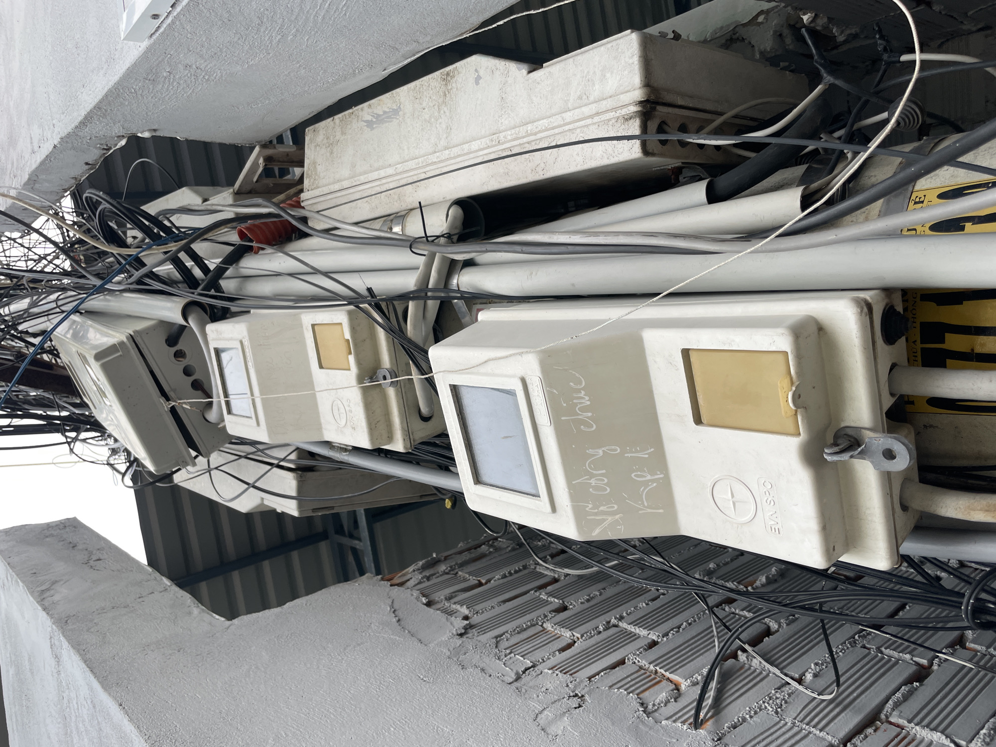 This is a low-angle, eye-level, medium shot of a cluster of electrical meters and a tangled mess of wires on a pole. The meters are white, rectangular boxes with some yellow flaps or panels. Some of the meters have writing on them, including what appears to be "EVN SPC". The surrounding environment includes a grey, rough-textured concrete wall on the left, partially covering a brick wall. On the right and in the background, there are more concrete structures and a corrugated metal roof. The sky is overcast and white. The overall scene suggests an urban outdoor environment, possibly in a residential or commercial area of Phu Thuy Ward, Vietnam, due to the "EVN SPC" marking, which refers to Electricity of Vietnam Southern Power Corporation. There are no people visible in the image. The focus is on the infrastructure and the chaotic arrangement of electrical components.