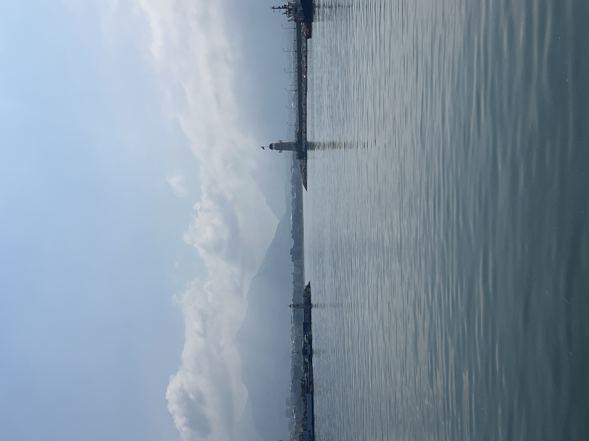 A serene coastal landscape, likely a harbor or bay, is depicted under a soft, partly cloudy sky. The image features a vast expanse of calm, deep blue-grey water in the foreground and midground, its surface gently rippled, reflecting the muted light.
Extending into the water from the left side of the frame is a long, dark pier or breakwater. A prominent white lighthouse with a dark top, possibly adorned with a red flag or cap, stands on this structure. Further along the pier, a larger vessel, appearing to be a cargo ship or ferry, is either docked or anchored.
In the distant background, a hazy city skyline can be discerned, stretching along the base of large, imposing mountains. These mountains are partially obscured by atmospheric haze and soft, white clouds that also dot the light blue sky. The overall lighting suggests it is daytime, likely late morning or early afternoon, with diffused light rather than direct sunlight.
No people are visible in the scene, and there is no discernible text. The tranquil composition emphasizes the natural beauty and the scale of the landscape. Given the context, the image captures a view of Alanya, Turkey, likely showcasing its harbor, the Mediterranean Sea, and the dramatic mountainous backdrop.