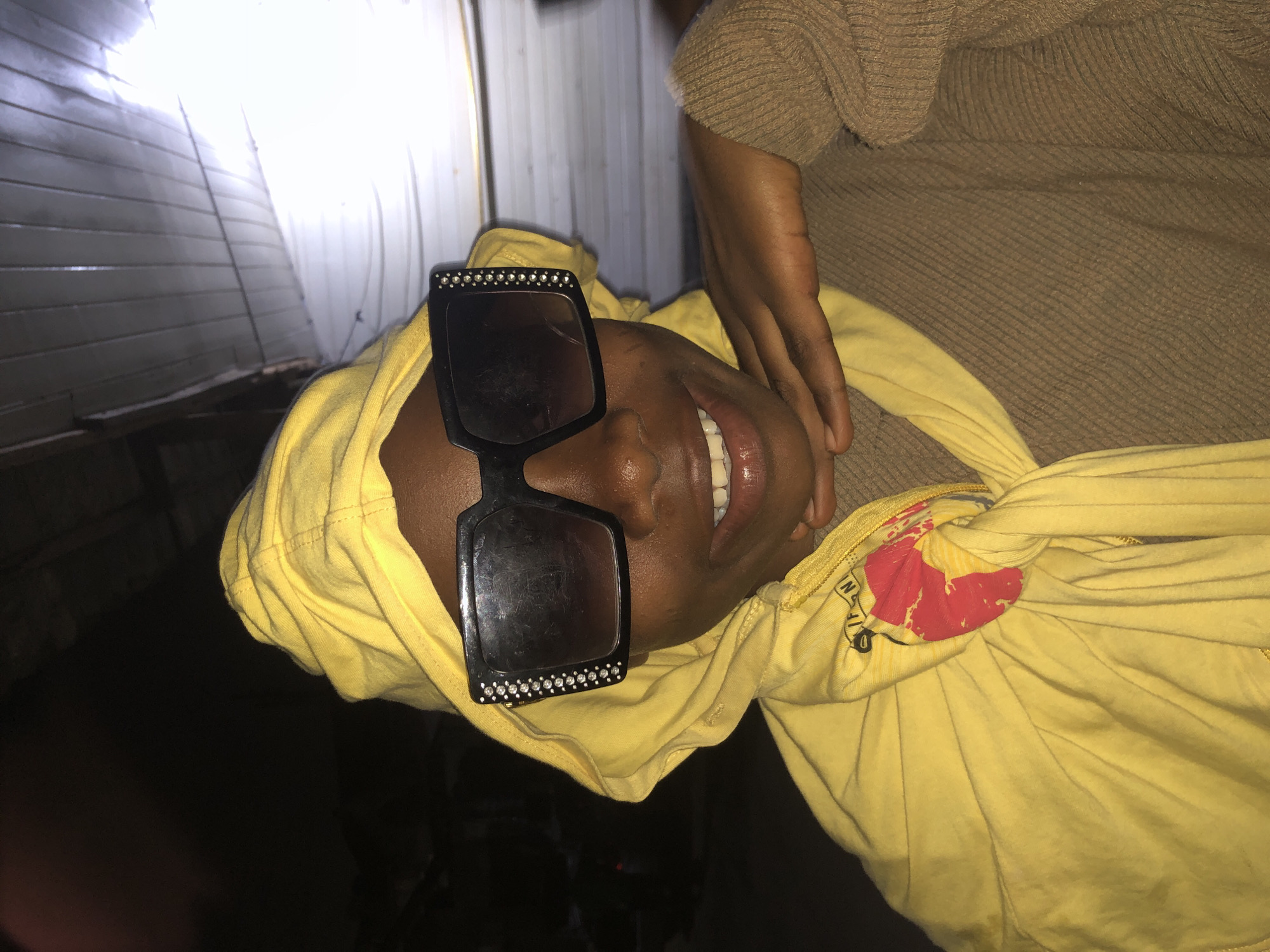 A close-up shot features a young Black individual from Ado, Nigeria, wearing a bright yellow hooded sweatshirt with the hood pulled up and large, square black sunglasses adorned with rhinestones. The individual is resting their chin on their right hand, with a slight smile showing their teeth. The background is dark, with horizontal planks visible at the top, suggesting an indoor or covered outdoor location. The lighting is dim, with a bright light source to the right, creating a somewhat dramatic effect. The individual's left shoulder is covered by a brown textured sweater. A red and yellow graphic is visible on the yellow sweatshirt.