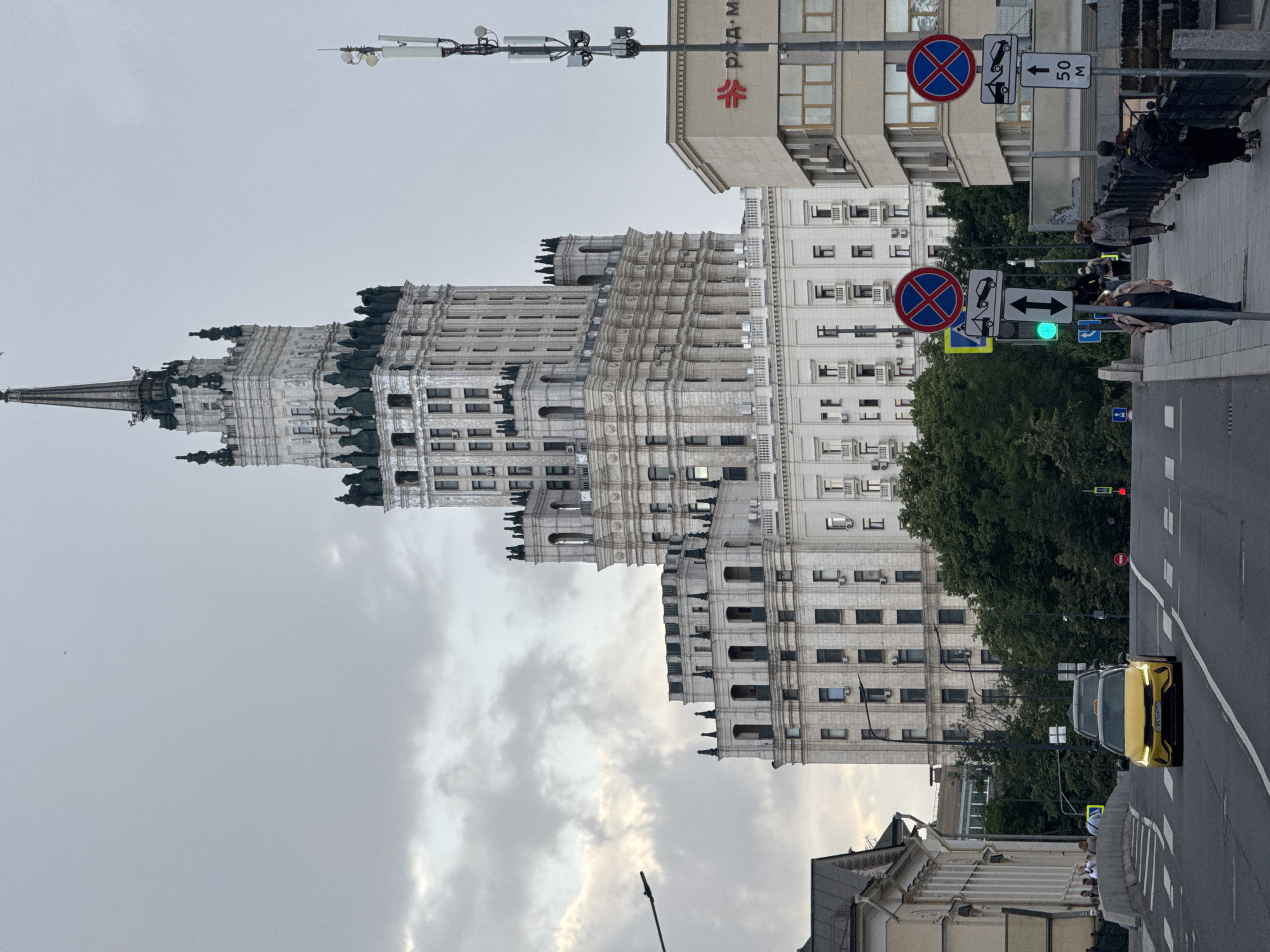 The image shows a view of the Kotelnicheskaya Embankment Building in Moscow, Russia, a prominent Stalinist skyscraper. The sky is overcast with dramatic clouds, and the lighting suggests it is either early morning or late afternoon. In the foreground, a yellow taxi is driving on a street with a pedestrian crossing. Several traffic signs are visible, including "no stopping" signs with a tow truck symbol. People are walking on a sidewalk to the right of the street, and a few individuals are visible further down the road near some buildings. The iconic skyscraper dominates the background, with its ornate architecture and spire. Trees and other buildings are also present in the scene, creating a typical urban environment.