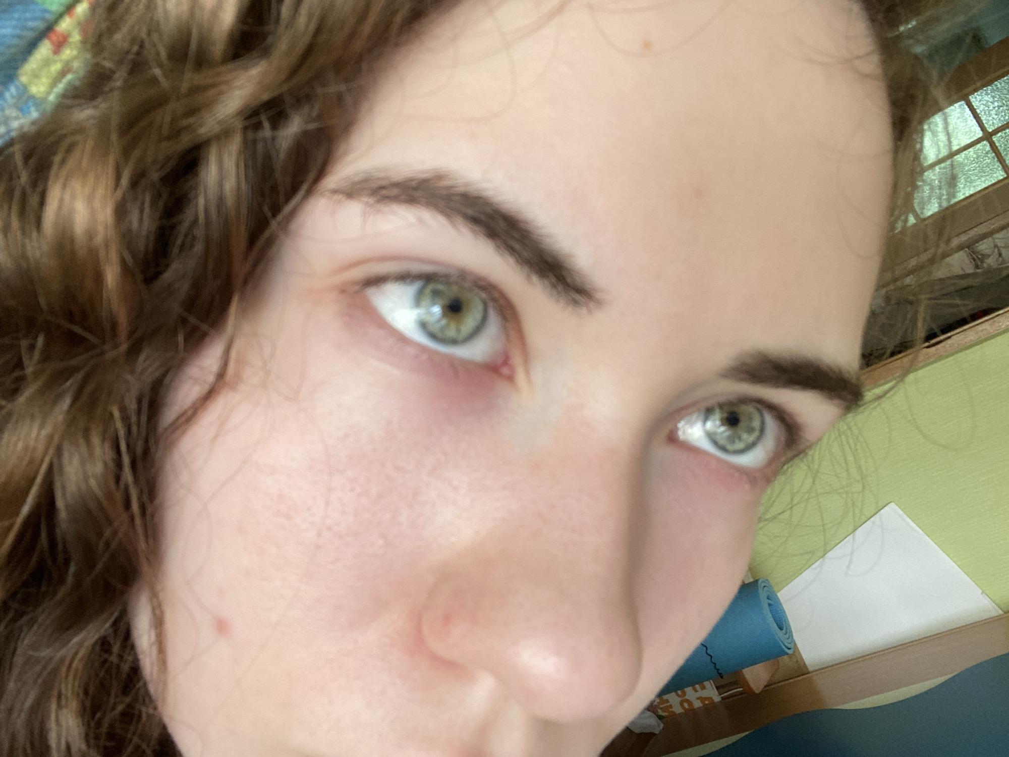 The image is a close-up, low-angle shot of a person's face, focusing on their eyes, nose, and cheek. The person has light skin, brown wavy hair, and striking green eyes with visible reflections. The background is out of focus but appears to be an indoor setting with a window showing greenery, a rolled-up blue mat, and wooden furniture. There is no visible indication of the city of Zavodoukovsk, Russia.