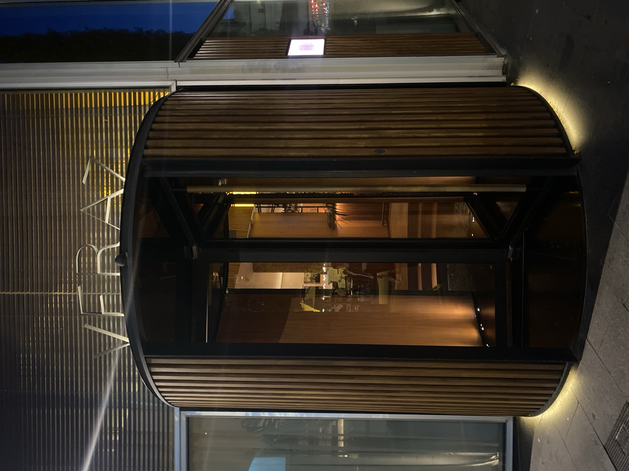 The image depicts the nighttime exterior entrance of Veraz Restaurant, located at The Barcelona EDITION hotel in Barcelona, Spain.
The central feature is a large, cylindrical revolving door constructed with dark vertical wooden slats and clear glass panels, set within a black metal frame. Above the entrance, the word "VERAZ" is displayed in illuminated, metallic letters against a background of horizontal blinds that cover a large window facade.
Warm yellow light emanates from beneath the base of the revolving door, illuminating the grey paved sidewalk directly in front of it. Through the glass panels of the revolving door, a warm-toned interior is visible, showing a staircase, wooden accents, and soft lighting. Faint, indistinct figures of people are discernible within the restaurant, suggesting activity.
To the left of the revolving door, a glass facade with more horizontal blinds is visible. To the right, another section of the building features a window reflecting ambient city lights and a portion of the dark blue night sky. A small, illuminated rectangular screen, possibly a digital display, is visible through this window. The overall scene is characterized by artificial lighting against the dark backdrop of a city night.