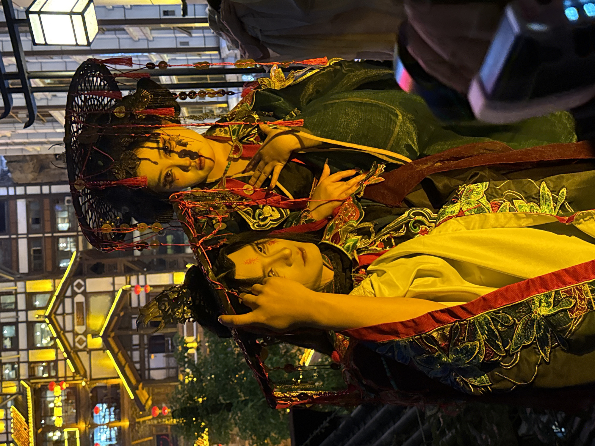 The image shows two people dressed in traditional Chinese clothing and makeup, likely performers or actors in a cultural performance or demonstration. They are in an outdoor setting, with the architecture visible in the background consistent with buildings in the city of Yuzhong District, China. The people are wearing elaborate headwear and clothing with intricate embroidery and ornamentation. The lighting suggests it may be evening or nighttime.