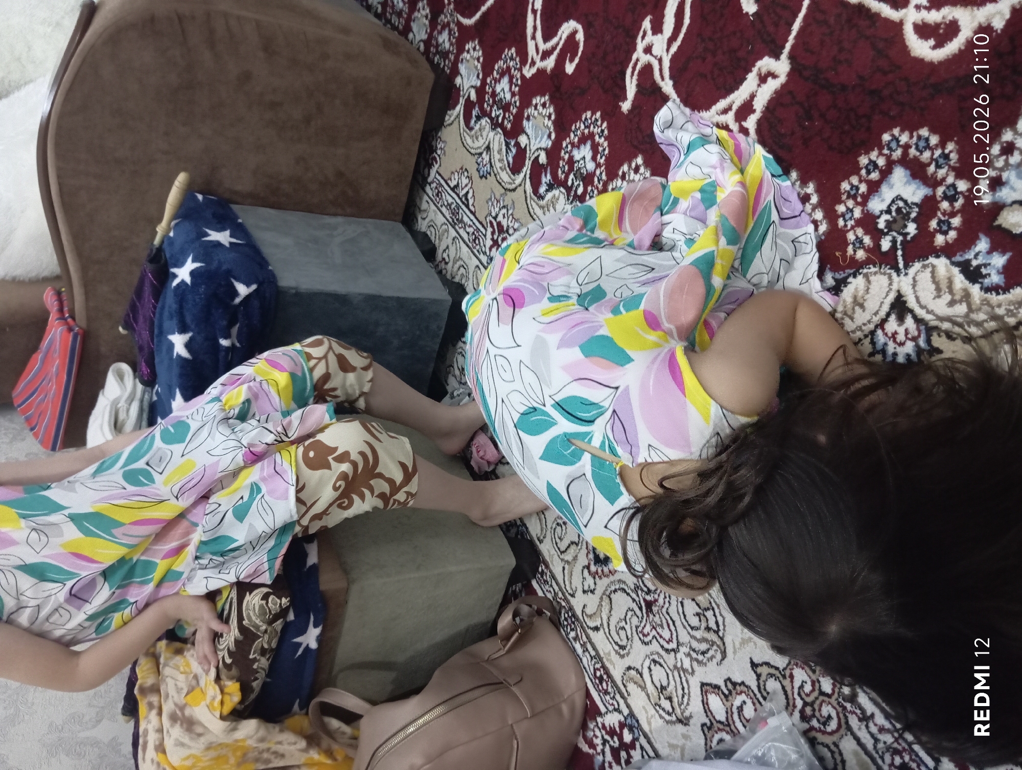 A prone female child with long dark hair is positioned on a carpet with ornate patterns, with her upper body on the carpet's patterned area, and her legs partially resting on what appear to be rectangular blocks. The child is wearing a floral-patterned pajama set. Nearby, there's a light brown handbag placed on the carpet. An armchair is visible in the upper-left corner. Several objects are placed on the blocks where the child's legs are positioned including a blue star patterned blanket. The photo was taken on May 19, 2026, at 21:10. The location is specified as Bekabad, Uzbekistan.