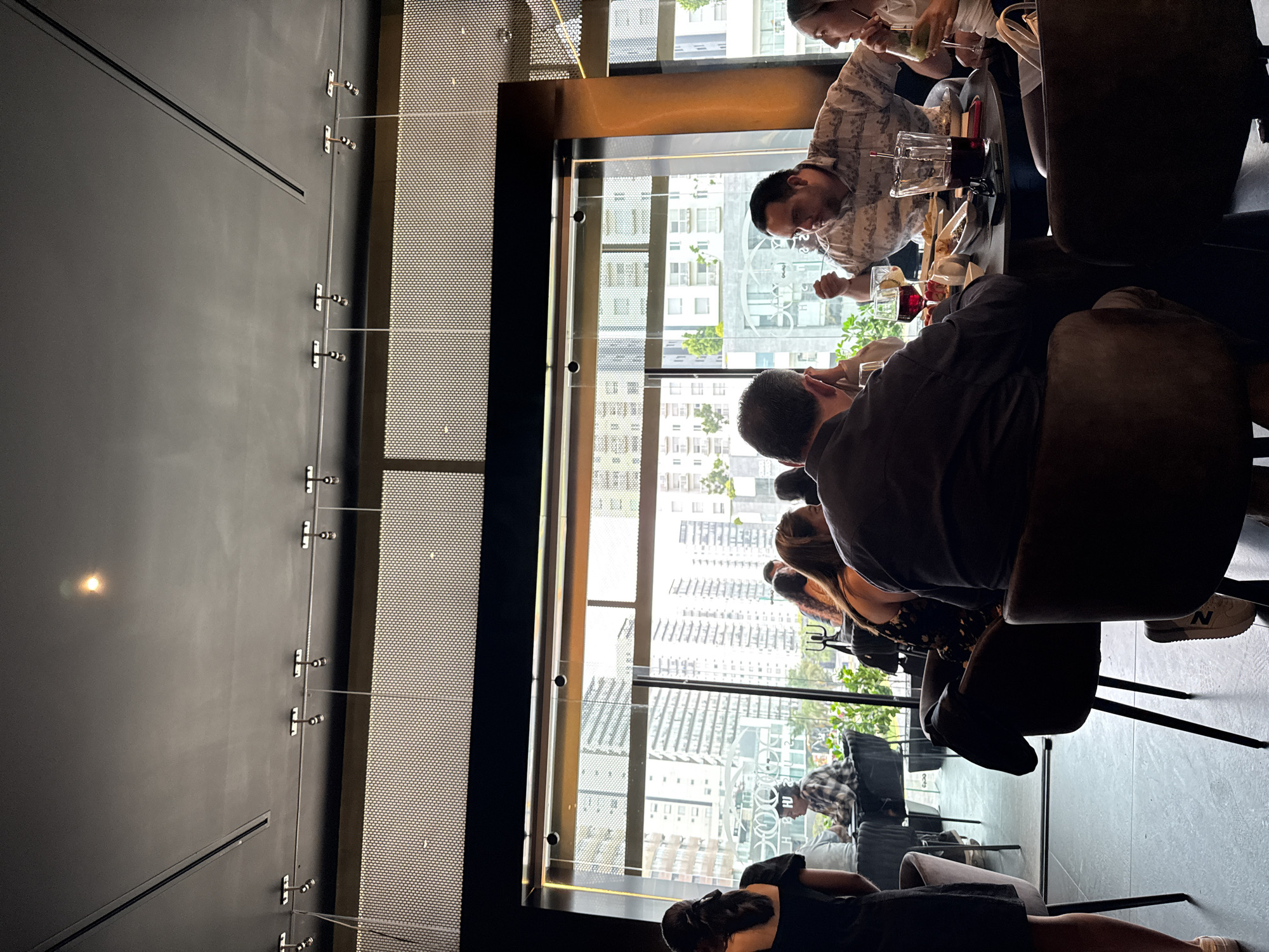 People are gathered inside a restaurant, seated at a table and engaging in conversation while enjoying drinks. The setting appears upscale, with a large window offering a view of urban architecture, likely a dense cityscape, outside. The restaurant has a modern design aesthetic, featuring a perforated screen on the left wall and sleek furnishings. The location appears to be in San Bernardino Tlaxcalancingo, Mexico.