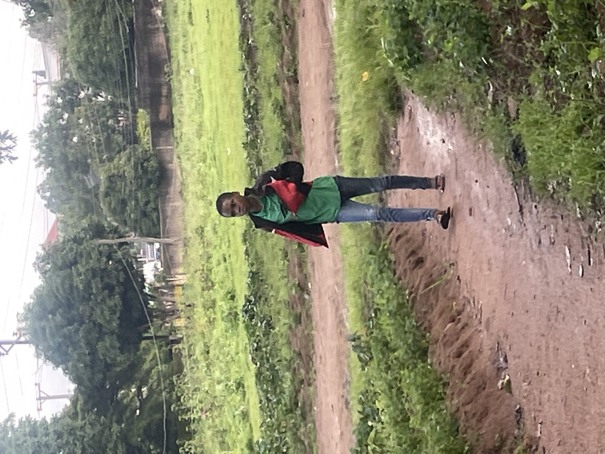 A young person walks on a dirt path through a grassy, somewhat overgrown area. The person is wearing a green t-shirt, blue jeans, and brown sandals. They have a red and black bag slung across their chest. Trees and some structures are visible in the background, suggesting an urban or semi-urban environment. The ground appears damp in places, indicating recent rain. The lighting is overcast, consistent with a cloudy day in Jos, Nigeria.