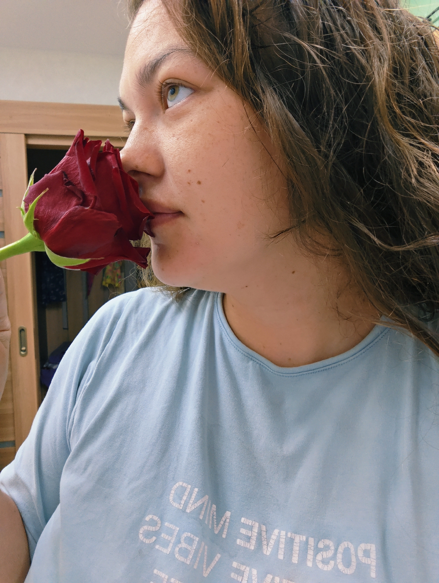 A woman is holding a dark red rose to her face, appearing to smell it. She has light skin with freckles and wavy brown hair. She is wearing a light blue t-shirt with white text that is upside down to the viewer. In the background, there is a wooden door.
The location is not deducible from the image. The city of Chaikovsky, Russia, cannot be determined.