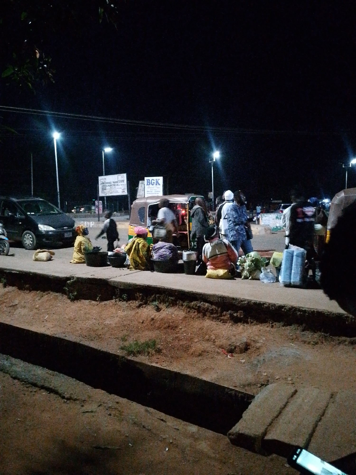 The image depicts a nighttime scene in Bukuru, Nigeria, likely a street market or roadside area. Several individuals are present, some appearing to be vendors and others customers or passersby. A three-wheeled auto-rickshaw, a common mode of transport and often used for vending in Nigeria, is visible, partially obscured by people and goods. Various items are laid out on the ground, suggesting a market setting; these include what appear to be produce like cabbage and other vegetables, as well as packaged goods and containers. A dark-colored car is parked on the left side of the image. The environment is characterized by a paved surface, a dirt embankment, and overhead streetlights illuminating the area. The overall impression is one of active commerce and daily life continuing after dark.