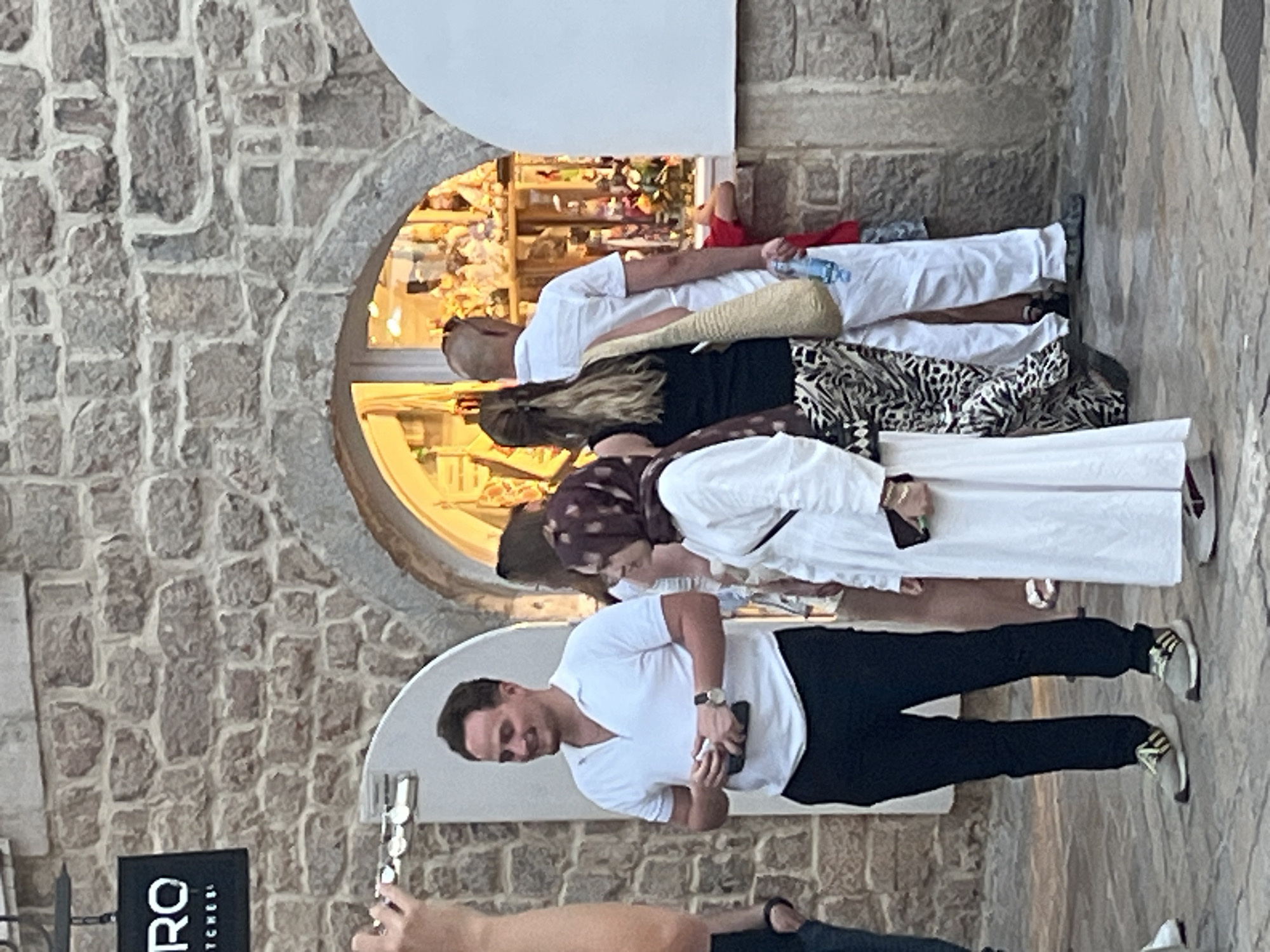 In Budva, Montenegro, a group of people stand near a stone building with an arched opening leading to a brightly lit shop window displaying merchandise. A man in a white polo shirt and black pants smiles as he holds a phone and a small white object. To his right, a woman in a white dress and a maroon hijab looks towards the shop. Behind them, a bald man in a white shirt and pants holds a bottle of water and a woven bag. Another woman with long blonde hair stands next to him. In the foreground, the legs of several individuals are visible, with one person wearing distinctive grey athletic shoes with yellow and black accents. The ground is paved with stone.
