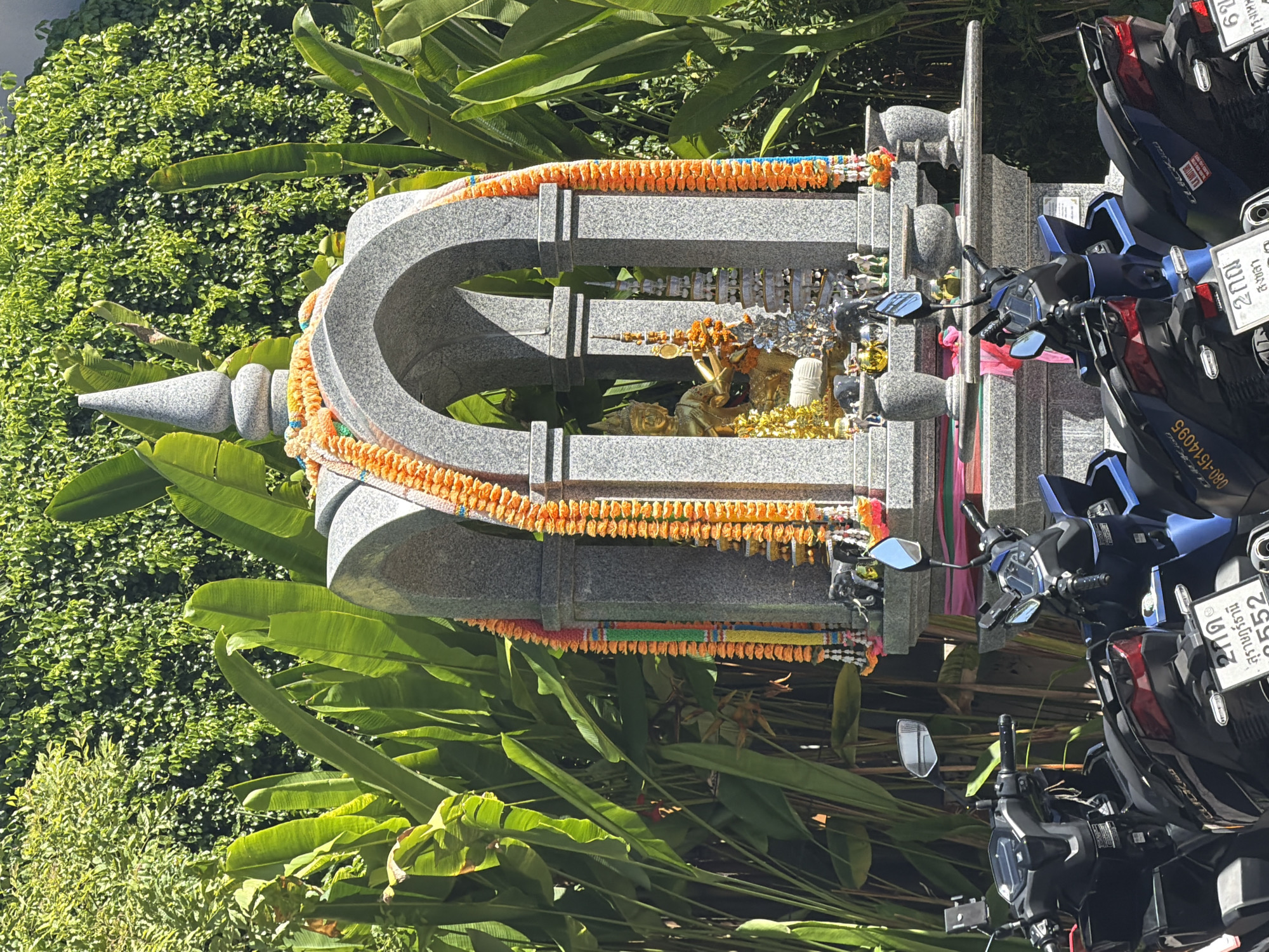 In Baan Chaweng, Thailand, a spirit house made of grey granite stands adorned with orange flower garlands and blue, green, and red ribbons. Within the spirit house, a golden statue of a seated figure, likely a deity, is visible, surrounded by smaller ornamental structures and figures, including elephants. The spirit house is situated amidst lush green foliage, with large, tropical leaves prominent in the background. In the foreground, the front portions of several parked motorcycles are visible, their handlebars and mirrors reflecting the bright sunlight. The scene appears to be outdoors, likely in a public area or near a residence, under a clear, sunny sky.