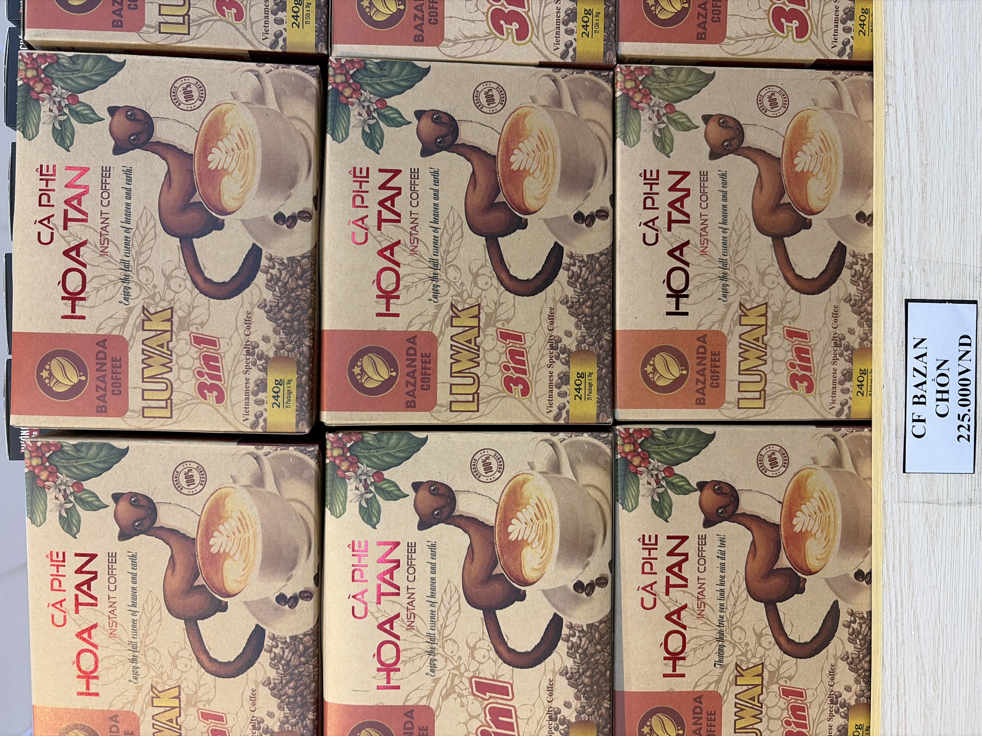 The image shows a retail display of coffee products in Đà Nẵng, Vietnam. The products are stacked in boxes with an illustrated design featuring a civet and a cup of coffee. The text on the boxes is in Vietnamese and English, indicating "CÀ PHÊ HÒA TAN INSTANT COFFEE" and "LUWAK 3in1 Specialty Coffee." The boxes are brown and orange, with red lettering. A price tag in the foreground reads "CF BAZAN CHÒN 225.000VNĐ". The lighting suggests an indoor setting, likely a store or market.