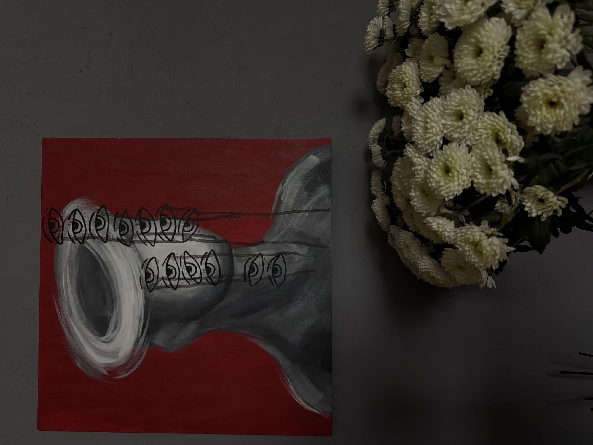 A painting on a red background hangs on a dark gray wall, to the right of which is a bouquet of white chrysanthemum flowers. The painting depicts a gray abstract shape with black outlines of eyes along the upper and middle sections. The background is solid red. The wall has a matte, slightly textured finish. The flowers are clustered together, with some green stems and leaves visible. The bottom right corner of the image is dark and partially obscured. There is no indication of the location being Ust-Ilimsk, Russia.