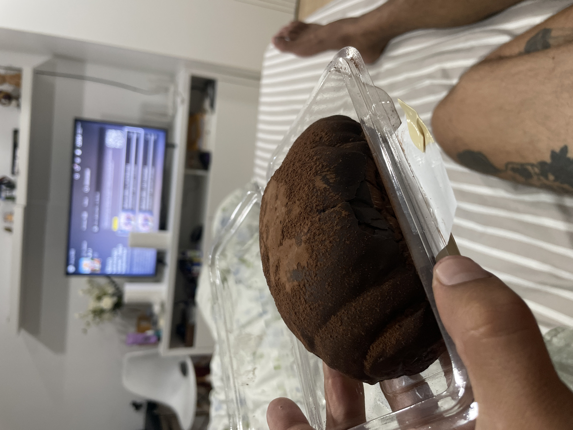 In a bedroom, a close-up shot focuses on a person's hand holding a plastic container with a dark brown, chocolate-dusted pastry inside. The pastry has a ridged surface and appears to be dusted with cocoa powder. The background is blurred, showing a white and grey striped bedspread. Further in the background, a white entertainment center with a television displaying a menu is visible. The television is positioned above a shelf containing various items. A white chair is also present in the room. The scene suggests a casual indoor setting, likely in a home.