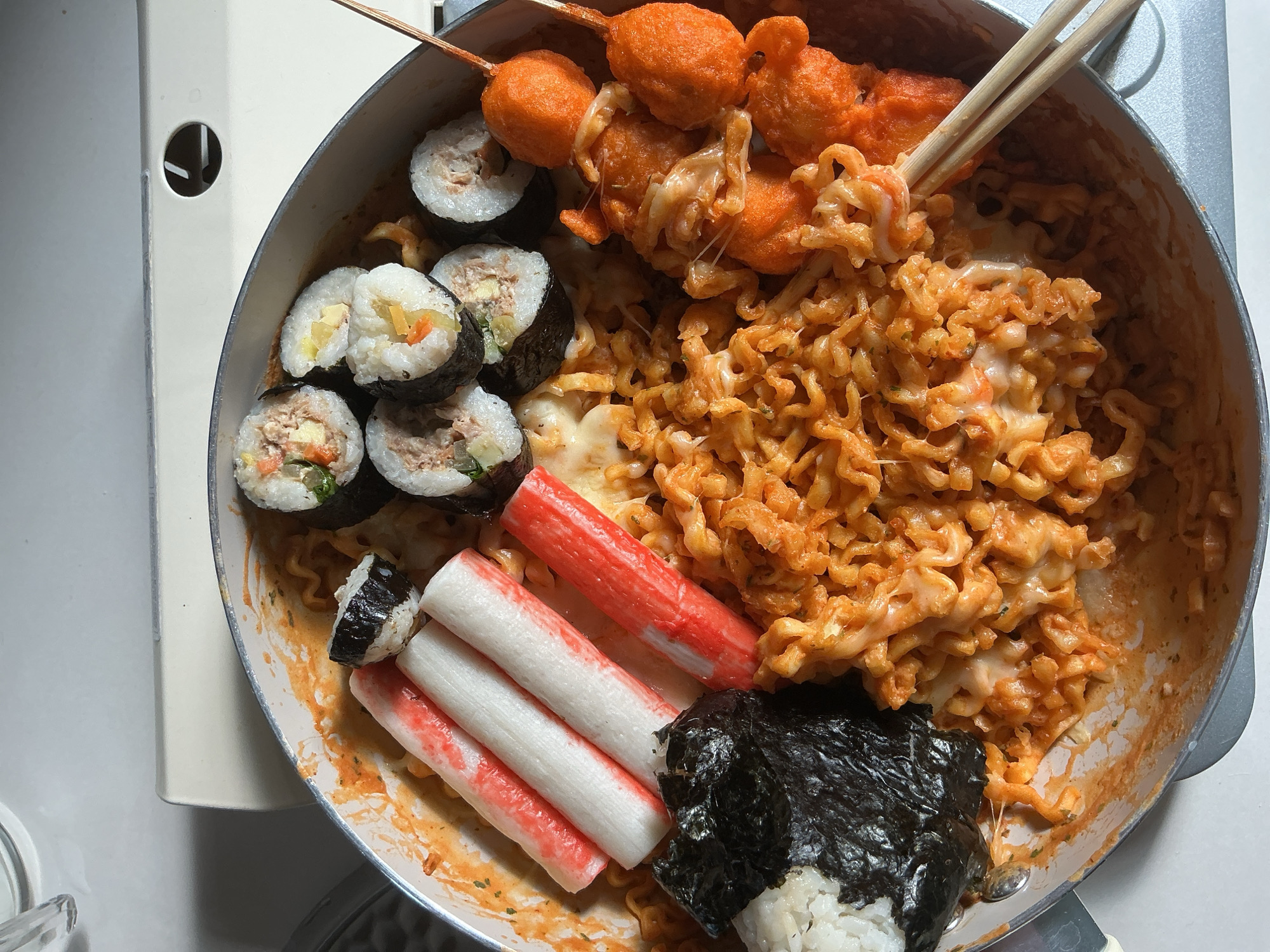The image shows a close-up, overhead view of a pot filled with a colorful assortment of food. At the center are tangled, bright orange noodles coated in a sauce and melted cheese. To the left, there are several white and red imitation crab sticks and a piece of sushi wrapped in nori seaweed with a side of white rice. To the right, a skewer holds several round, bright orange pieces of what appear to be fried fish cakes, also covered in melted cheese. The food is arranged in a shallow, grey-rimmed pot. The background is a plain white surface, suggesting the food is placed on a table or counter. There are no people or any discernible landmarks within the image to indicate a specific location in Davao City, Philippines.