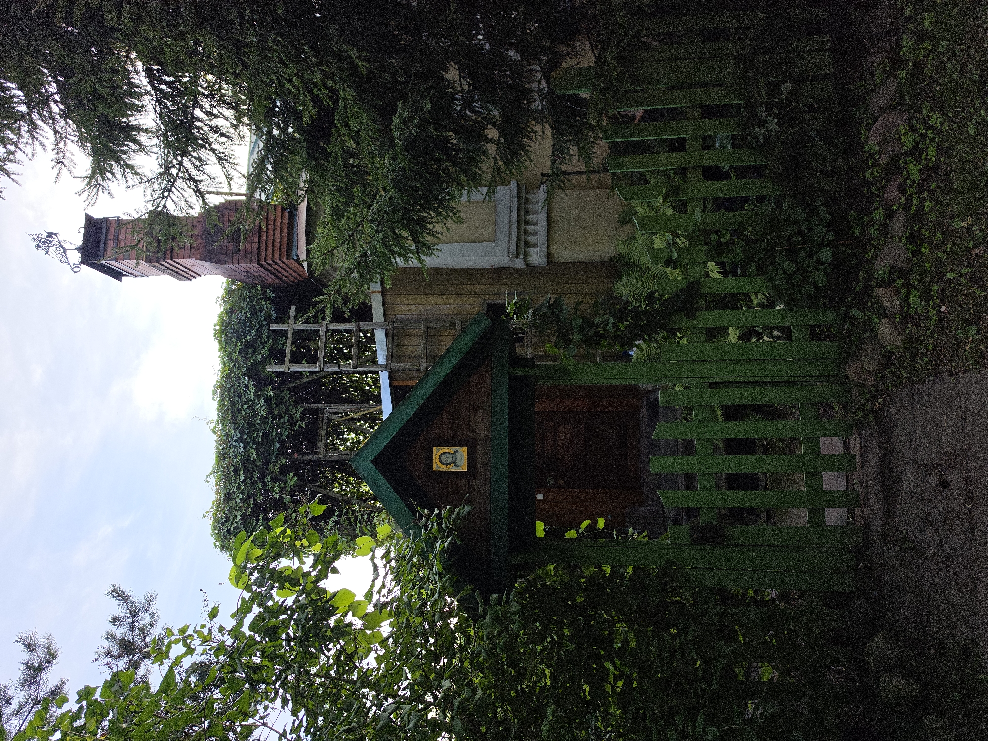 The image shows a modest wooden structure with a green, gabled roof, resembling a small outbuilding or entrance to a property. Above the dark wooden door of this structure, an icon featuring a bearded man with a halo is affixed. The entrance is enclosed by a low, green wooden picket fence. To the left and right of the entrance, lush green foliage and trees are prominent, partially obscuring the view. In the background, a red brick chimney with a metal weather vane is visible, along with a portion of a building with light-colored walls and decorative architectural elements. The scene appears to be in a residential area, likely Strelna, Russia, given its rustic and natural elements. The lighting suggests it is daytime, possibly late afternoon or early evening, with soft, diffused light.