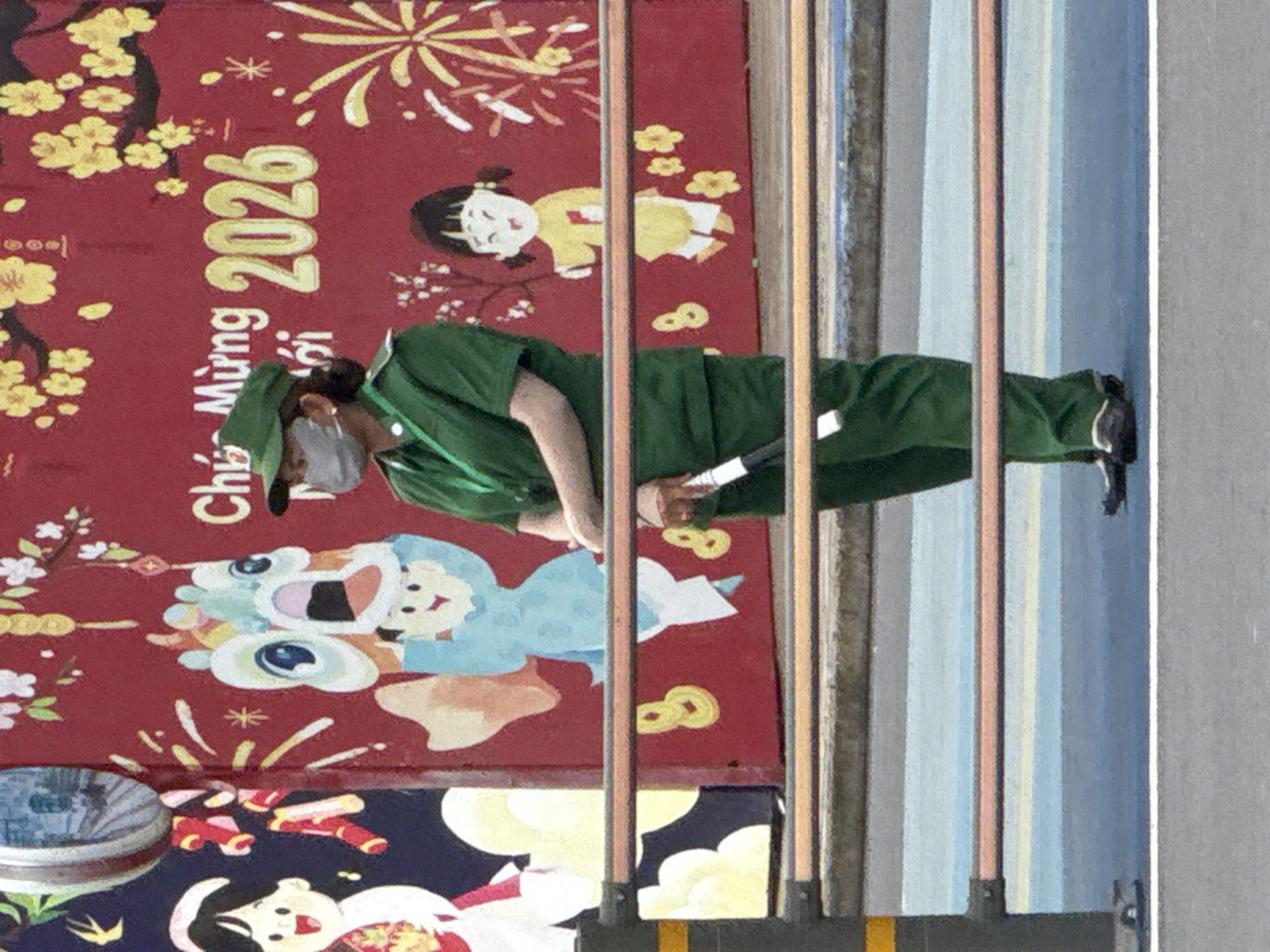 A full-shot, eye-level medium shot photograph captures a Vietnamese woman in a green uniform and cap standing outdoors. She wears a gray face mask and holds a white baton in her right hand. The woman is positioned in front of a mural decorated with cartoon figures, stylized flowers, and the text "Chúc Mừng 2026" in yellow lettering against a red background. The mural is visible through a horizontal railing in the foreground. The woman's dark hair is tied back. Her uniform consists of a short-sleeved green shirt and green trousers. She wears dark shoes. The background mural depicts a playful scene with a dragon head, children, and festive elements, characteristic of Tet (Vietnamese Lunar New Year) decorations. The overall setting appears to be a public space, possibly a street or plaza in Thủ Đức City, Vietnam, given the thematic elements suggestive of a holiday celebration. The lighting is consistent with daylight, casting minimal shadows.