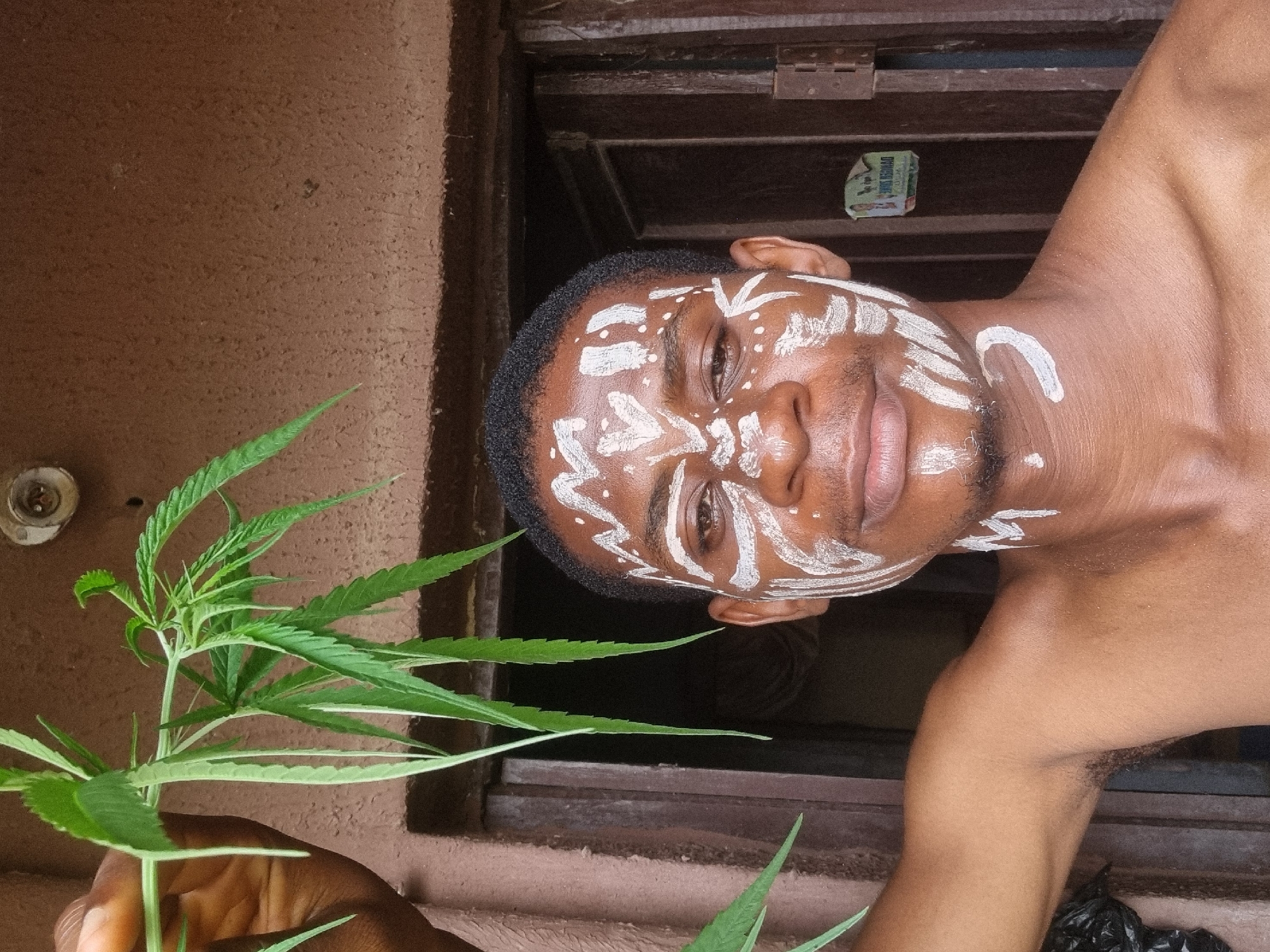 A partially nude African man with white paint markings on his face and neck holds a cannabis plant. He stands in front of a window. The background shows a brown, textured wall. The location is Onitsha, Nigeria.