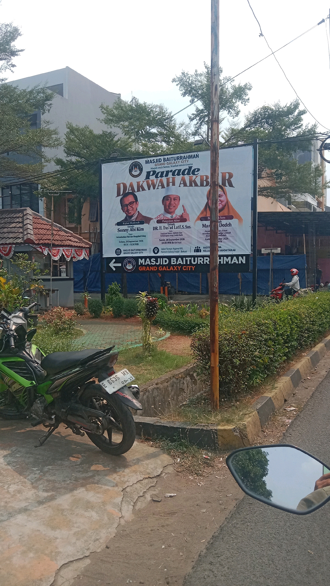 A horizontal, medium shot captures an outdoor scene in front of Masjid Baiturrahman Grand Galaxy City, Bekasi, Indonesia. The foreground features a portion of a green and black motorcycle parked on a cracked concrete surface. To the right, a dark asphalt road is visible, reflecting a cloudy sky and trees in a motorcycle's rearview mirror. Beyond the motorcycle, a well-maintained garden with green bushes and scattered dry grass lines a curb with yellow and black markings. A vertical wooden pole stands prominently in the middle ground.
Dominating the background is a large white banner for "MASJID BAITURRAHMAN GRAND GALAXY CITY." The banner advertises a "DAKWAH AKBAR" event, featuring portraits and details of three speakers: Ustadz Sonny Abi Kim, DR. H. Da'ad Latif, S.Sos., and Mariam Deddeh. Text on the banner indicates event dates and times: "Selasa, 23 September 2026 09.30 - 11.30 WIB" and "Senin, 28 September 2026 19.00 WIB." An arrow points left towards the mosque entrance, with the mosque's name and location "Jalan Taman Anggrek VIII, Taman Galaxy Indah, Bekasi" also displayed. To the left of the banner, a building with red-tiled roofing and white walls is partially visible, with a small red and white decorative structure in front of it. Trees with lush green foliage line the top of the banner and extend beyond it. In the far right background, a person wearing a red helmet is riding a red motorcycle on the road.