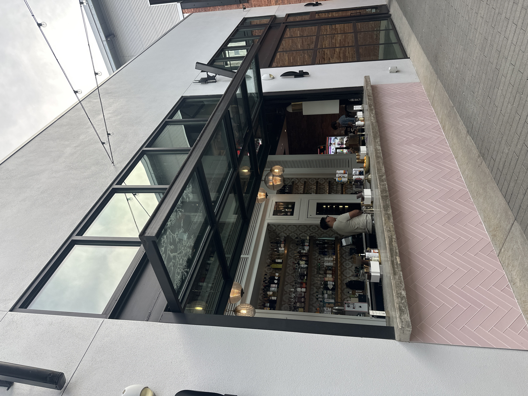 Here's a concise description of the image:
The image presents an outdoor bar area, seemingly a restaurant extension, in what appears to be a modern, possibly gentrified area. A multi-paneled window is partially open, revealing the interior of the bar with shelves of alcohol bottles. A bartender is visible, positioned behind the counter. The exterior of the building is predominantly white, with some exposed brick. The outdoor counter is bordered by pink herringbone-patterned tiles. String lights are visible along the building's edge. The location is likely in Tampa, Florida, based on the prompt.