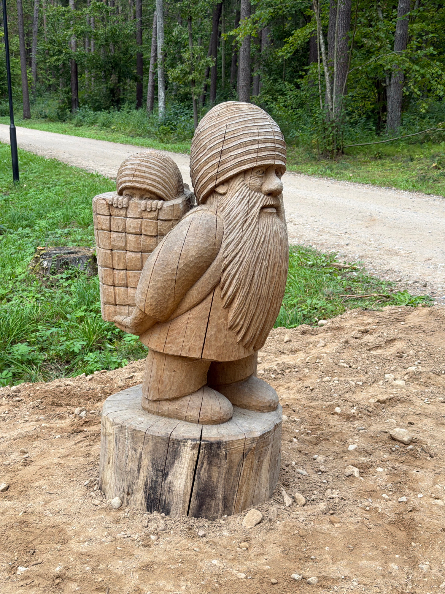 The image depicts a wooden sculpture of a gnome carrying a child in a basket on its back, situated in the Tērvete Nature Park in Tērvete, Latvia. The sculpture is carved from wood and mounted on a tree stump. The gnome has a long beard and is wearing a striped hat. The child's face is peeking out from the basket. The sculpture is placed on sandy ground with small stones. In the background, there is a gravel path winding through a grassy area and a dense forest with tall trees. A black lamppost is visible to the left of the path. The overall environment is natural and rustic.