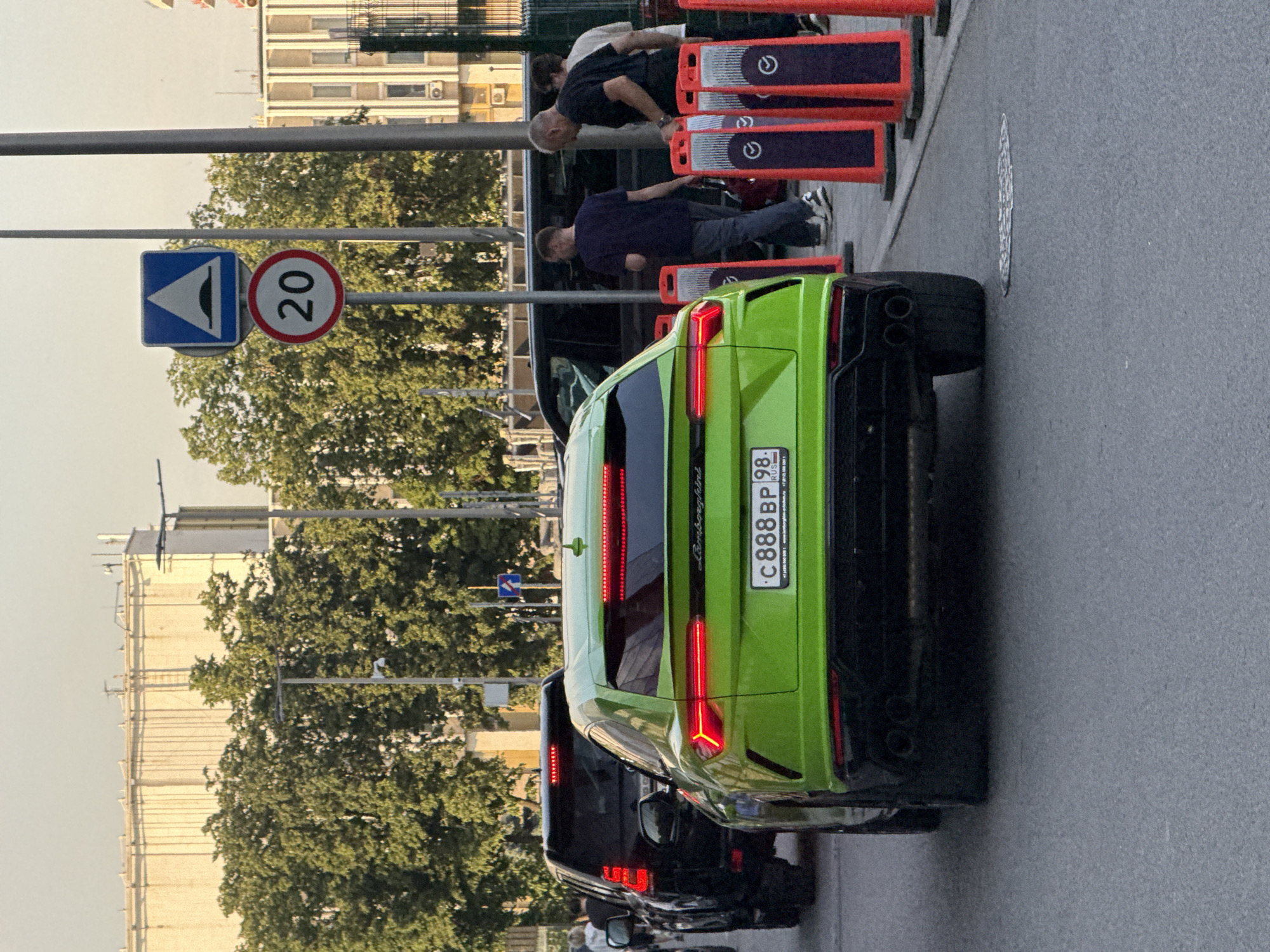 The image depicts a street scene in Moscow, Russia, with a bright green Lamborghini Urus as the primary subject, parked behind a black SUV. The car's distinctive LED taillights are illuminated. Behind the vehicles, several people are visible, including pedestrians and individuals near a set of orange traffic cones. A speed limit sign indicating "20" and a warning sign for a speed bump are also present. The background features trees and buildings, suggesting an urban environment. The lighting indicates it is daytime, likely late afternoon or early evening.