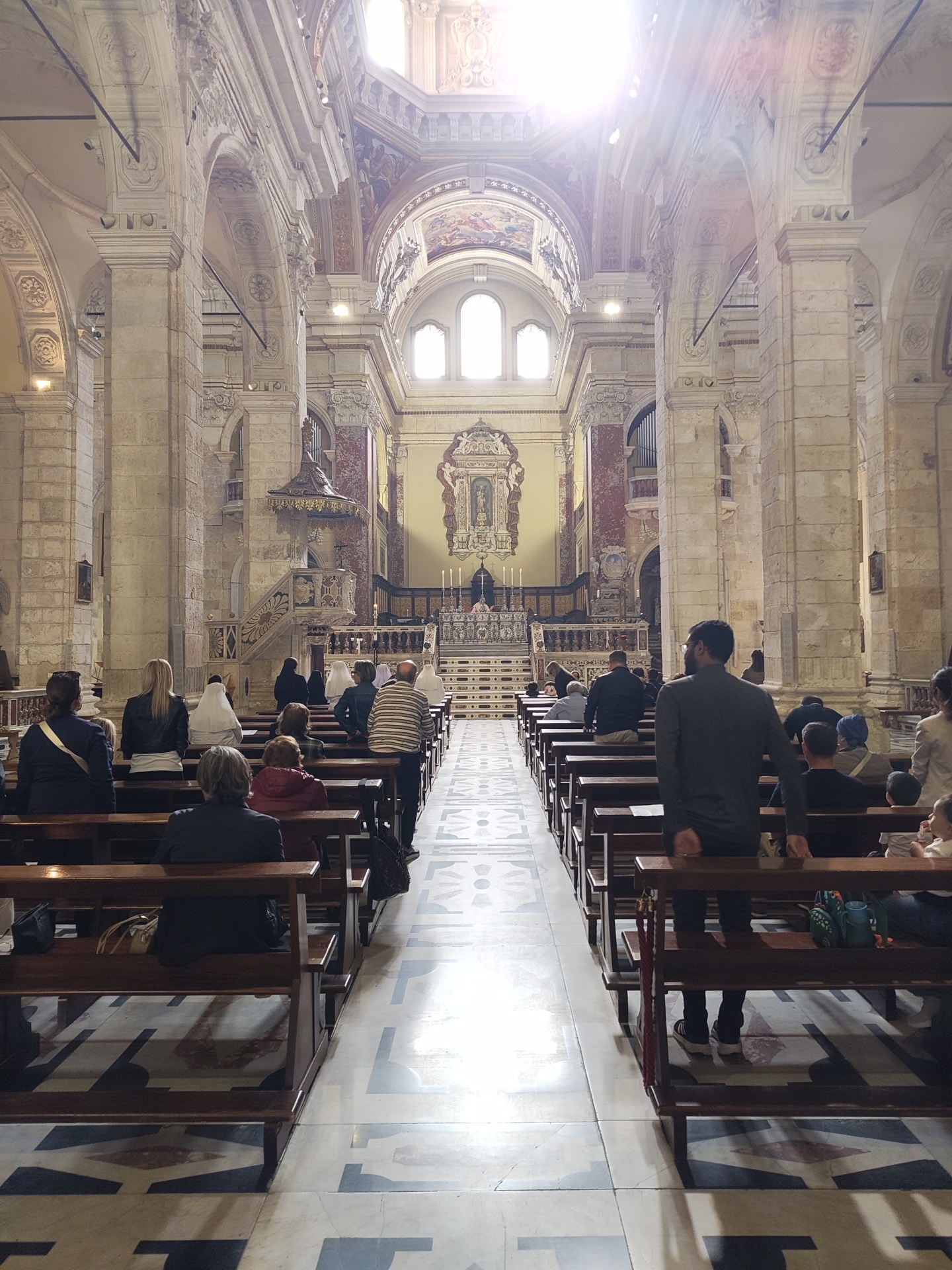 The image shows the interior of the Cagliari Cathedral in Cagliari, Italy. The nave is lined with pews filled with people attending a service. Tall, ornate columns support arches that lead to the dome. Sunlight streams in from windows at the front of the cathedral. The floor is patterned with geometric designs. In the background is the altar area with candles and religious artifacts. A pulpit is visible to the left. The people are of various ages and are dressed in everyday clothing.