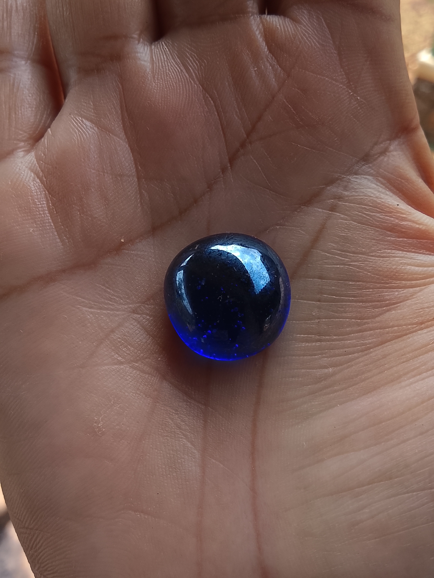 A close-up shot shows a hand holding a dark blue glass marble in Dawung, Indonesia. The marble, which has small specks of lighter blue within it, rests in the palm of a person's hand. The hand displays distinct lines and creases, indicative of natural skin texture. The background is blurred, but appears to be outdoors with a reddish-brown hue.