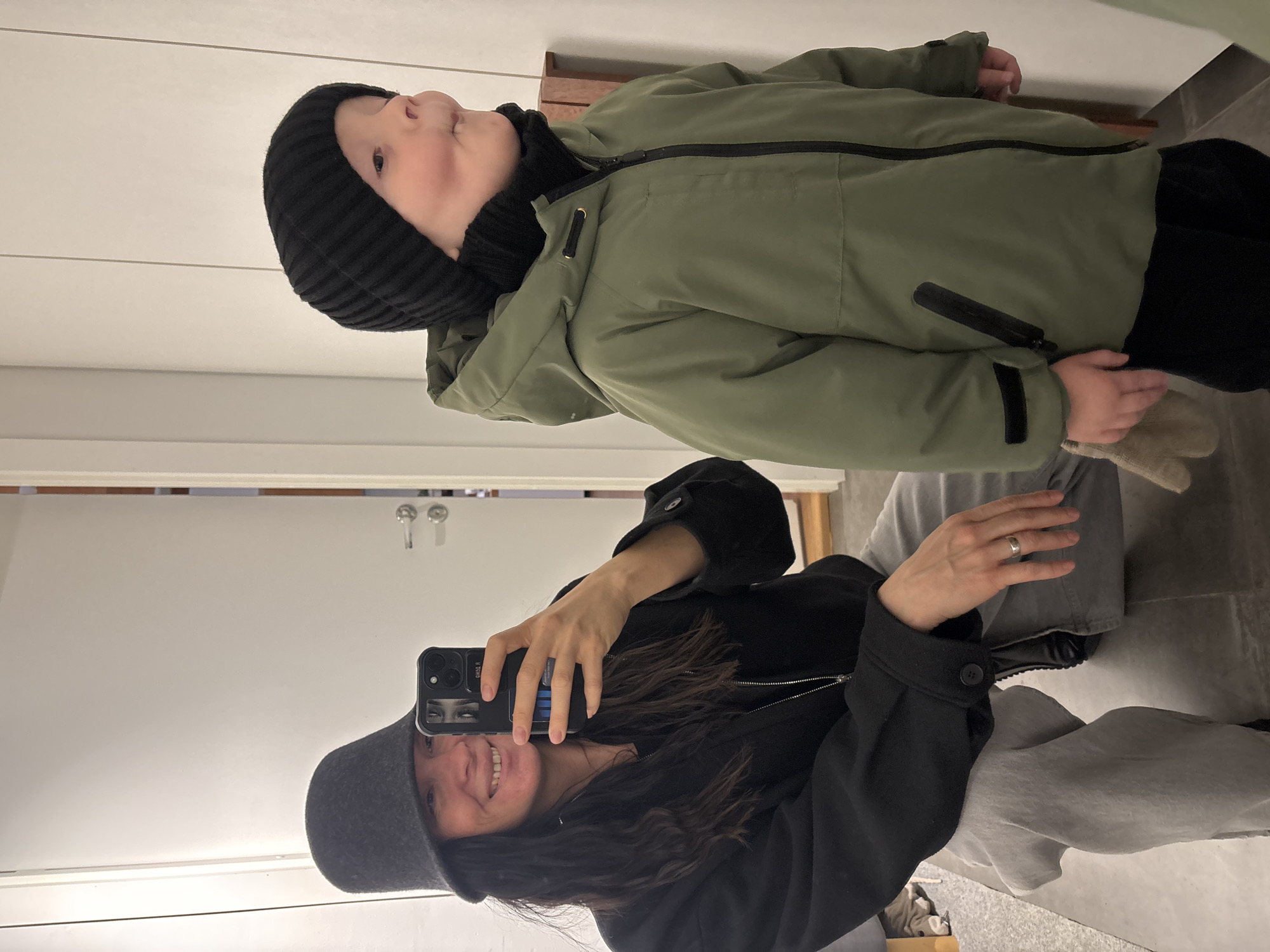 A person with long, dark hair and wearing a gray hat and black jacket holds a smartphone in a mirror reflection, taking a photo of themselves and a child. The child, wearing a black knit hat and a green winter coat, is positioned slightly above and to the left of the person holding the phone. The environment appears to be an indoor setting, possibly a hallway, with a white door visible in the background. The location is Chaikovsky, Russia.