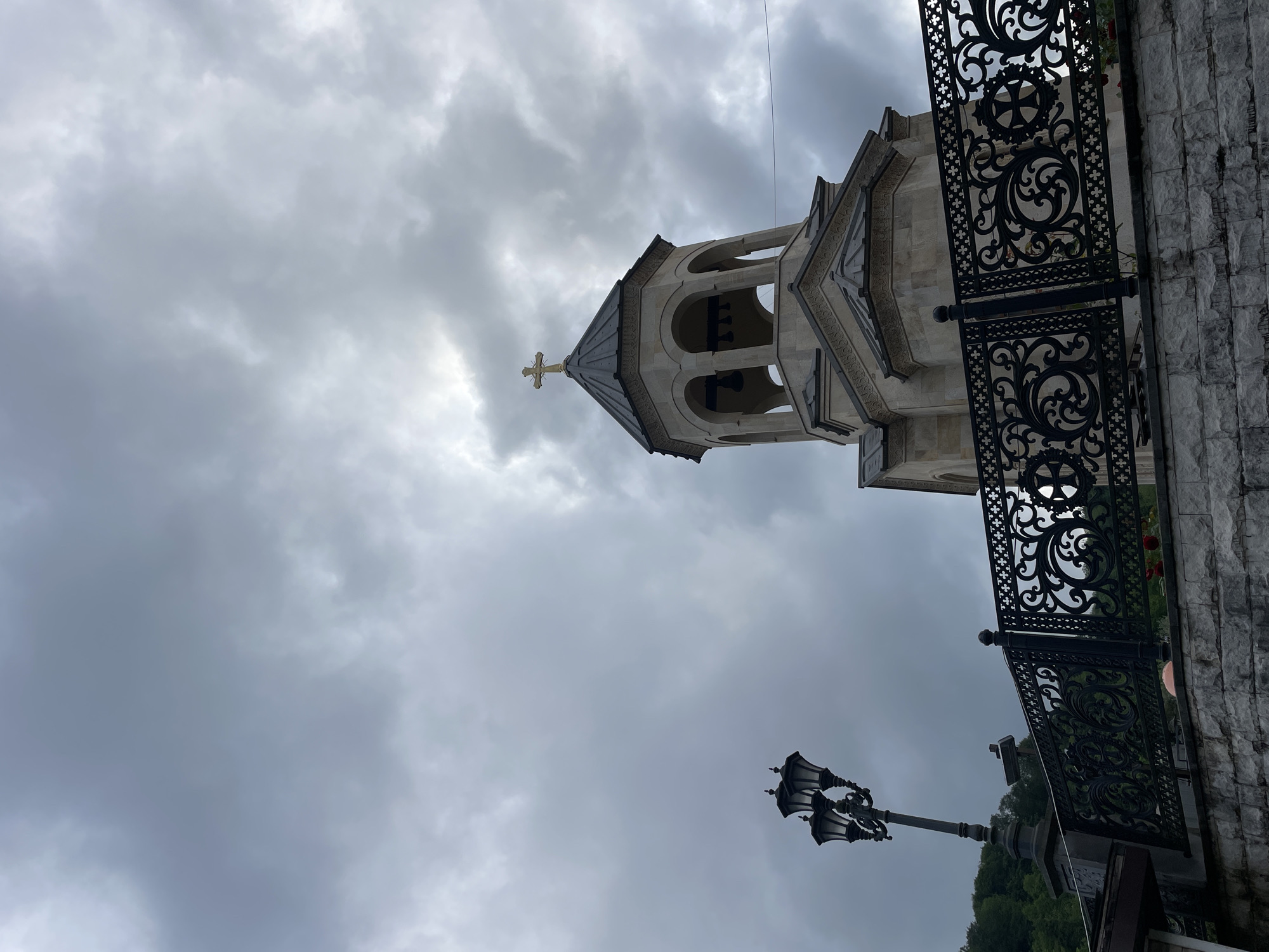 The image shows a church tower in Sochi, Russia, against a cloudy sky. A gold cross sits atop the tower. A decorative black metal fence is visible on the right. A black lamppost stands in the lower portion of the image.