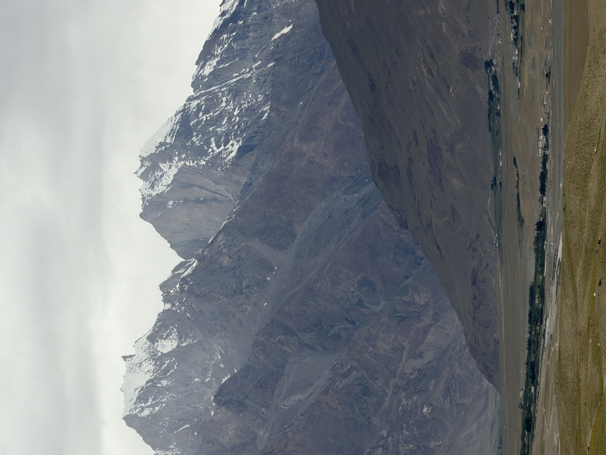 A mountainous landscape is captured in the image, with snow-capped peaks in the background. The foreground features a wide, arid valley with sparse vegetation. A small village is visible in the middle ground, with a cluster of white buildings and some scattered trees. The sky is overcast, contributing to the muted tones of the scene. The location is identified as Pibiting, India. There are no people or animals clearly visible in the image.