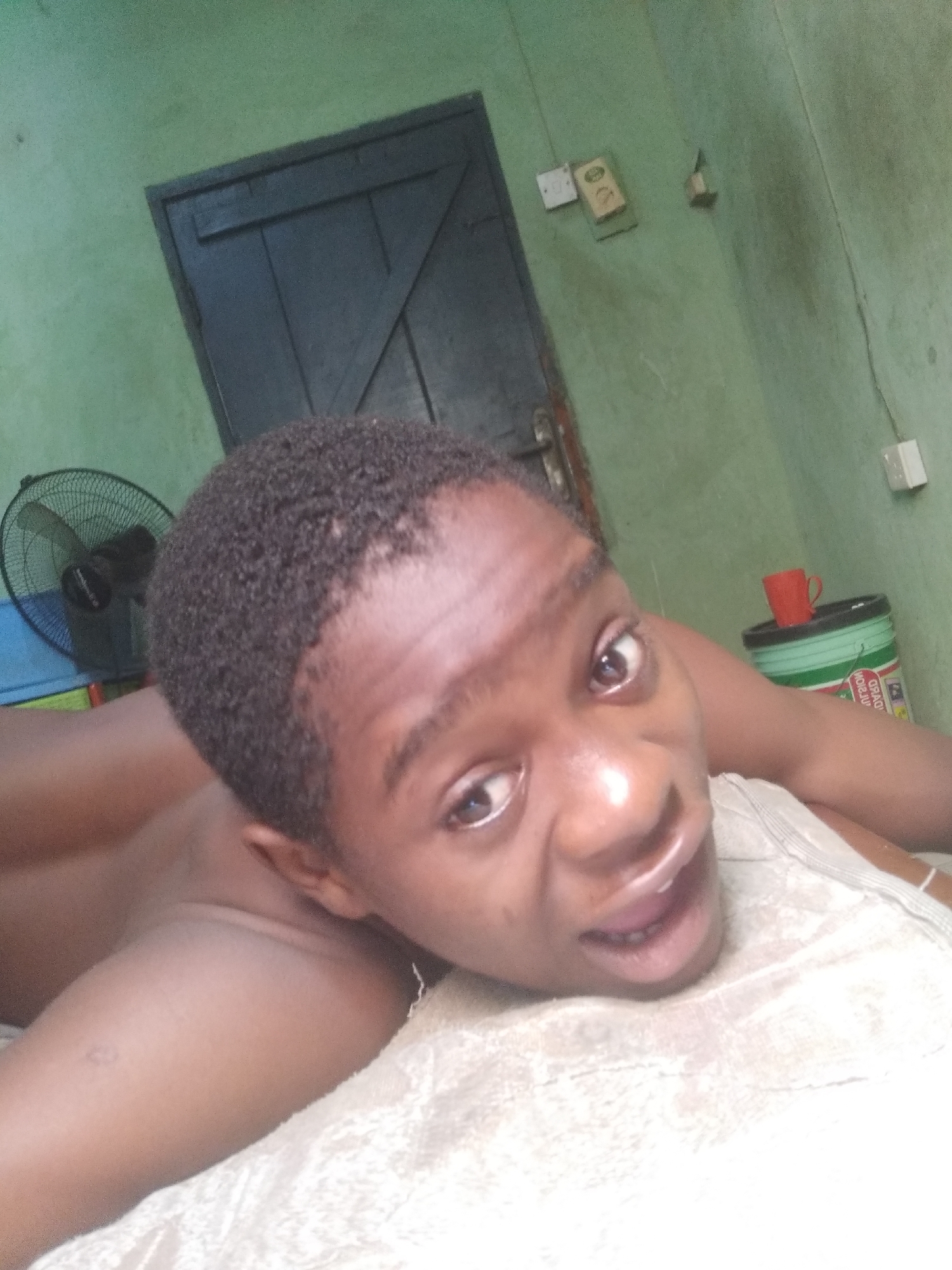 A person with short, dark hair is lying on a light-colored textured surface, possibly a bed, with their head tilted towards the camera. Their mouth is slightly open, and they are looking directly at the camera.
In the background, there is a dark-colored door, a light-green wall, and various objects, including electrical outlets and a cylindrical container with a red mug on top. A black fan is visible on the left side of the frame.
The image was taken indoors, possibly in Shangisha, Nigeria.
