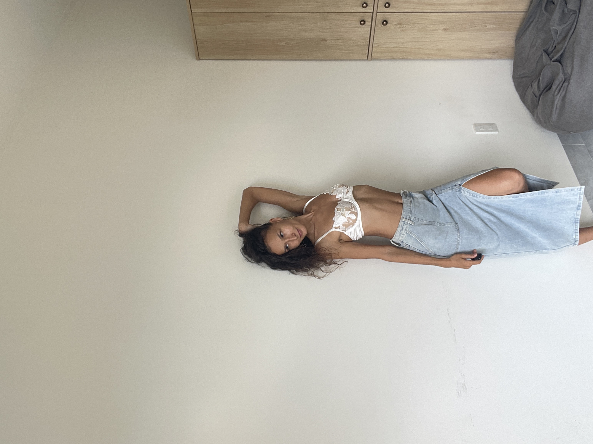 A woman with dark, curly hair stands leaning against a white wall in a room. She is wearing a white lace bralette and a light blue denim maxi skirt with a high slit. Her left arm is raised, with her hand behind her head, and she is looking to her right. A wooden cabinet with two doors is visible to her right, and a gray beanbag chair is on the floor to the right of the cabinet. There is an electrical outlet on the wall behind her. The scene is indoors and appears to be a residential setting. The location of Phu Thuy Ward, Vietnam is not determinable from the image.