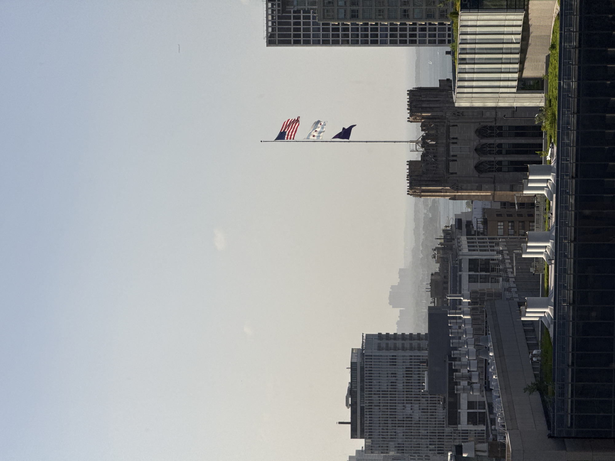 A cityscape view shows various buildings against a hazy sky. In the foreground, a green roof with prominent ventilation pipes is visible on top of a modern building. Behind this, a Gothic-style building with arched windows stands tall. To the right, a contemporary glass and metal building with vertical elements is prominent. Further in the background, a cluster of flags, including the American flag and the Chicago flag, flies from a flagpole. To the left, a large, multi-story building with a grid-like facade dominates the scene. In the far distance, silhouetted buildings are visible through the haze, suggesting a larger urban area. A faint airplane trail is discernible in the upper right portion of the sky. The scene appears to be an aerial view of Chicago, given the presence of the Chicago flag and the characteristic architecture.