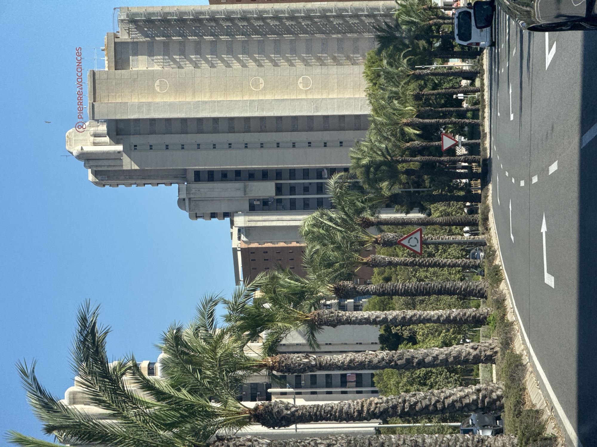 The image shows a sunny day in Benidorm, Spain, with a view of a wide road lined with palm trees. In the background, there are several tall, modern buildings, some of which are concrete-colored and feature repetitive window patterns. One prominent building has a red sign at the top that reads "PIERRE VACANCES". The road has white lane markings, indicating multiple lanes of traffic. A white van is visible in the right lane, and a dark car is partially visible in the foreground, suggesting the image was taken from a moving vehicle. Traffic signs, including a triangular sign with a circular arrow, are also present along the roadside. The overall environment appears to be a developed urban area with a clear blue sky overhead.
