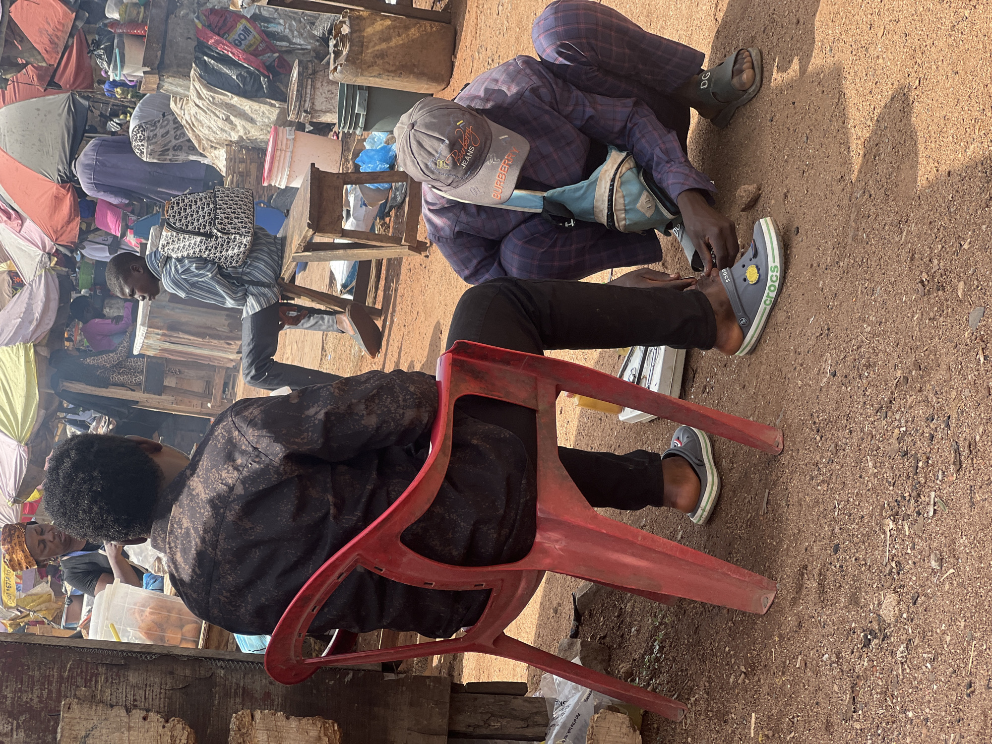 The image depicts a vibrant outdoor market scene in Gura Top, Nigeria, under bright daylight. In the foreground, two men are engaged in an interaction. One man is seated on a red plastic chair, viewed from behind, wearing a dark patterned long-sleeved shirt and black trousers. His short, curly hair is visible. Facing him, another man is squatting, appearing to adjust something on the seated man's foot or shoe. This man wears a plaid long-sleeved shirt, dark plaid trousers, and a grey cap featuring "JEANS" and "Burberry" text. He also carries a light blue sling bag.
He is seen holding a grey Crocs shoe with a white and green stripe and a yellow emblem, clearly displaying the word "crocs" on its side. The seated man's visible foot wears a dark sandal with "DG" written on it. The ground is a dusty, stony dirt surface, with strong shadows indicating a sunny day.
The background reveals a bustling market environment with various makeshift stalls, colorful umbrellas, buckets, and bags. Other individuals, including one wearing a striped shirt and carrying a bag resembling a Dior pattern, are partially visible amidst the market activity. Additional text discernible in the scene includes "GOM" and partial phrases like "Dial 671 to" and "Part of the bulk, not t" on plastic packaging.