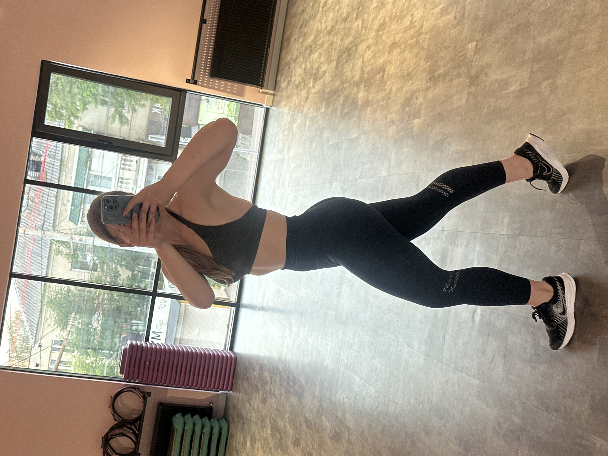 A woman is taking a mirror selfie in a gym. She is wearing a black sports bra and black leggings, and black sneakers with white soles. She is holding a smartphone in her hands to take the picture. The gym has a large window that looks out onto buildings and trees. There are exercise equipment such as stacked aerobic steps and exercise hoops visible in the background. The floor is made of gray tiles.