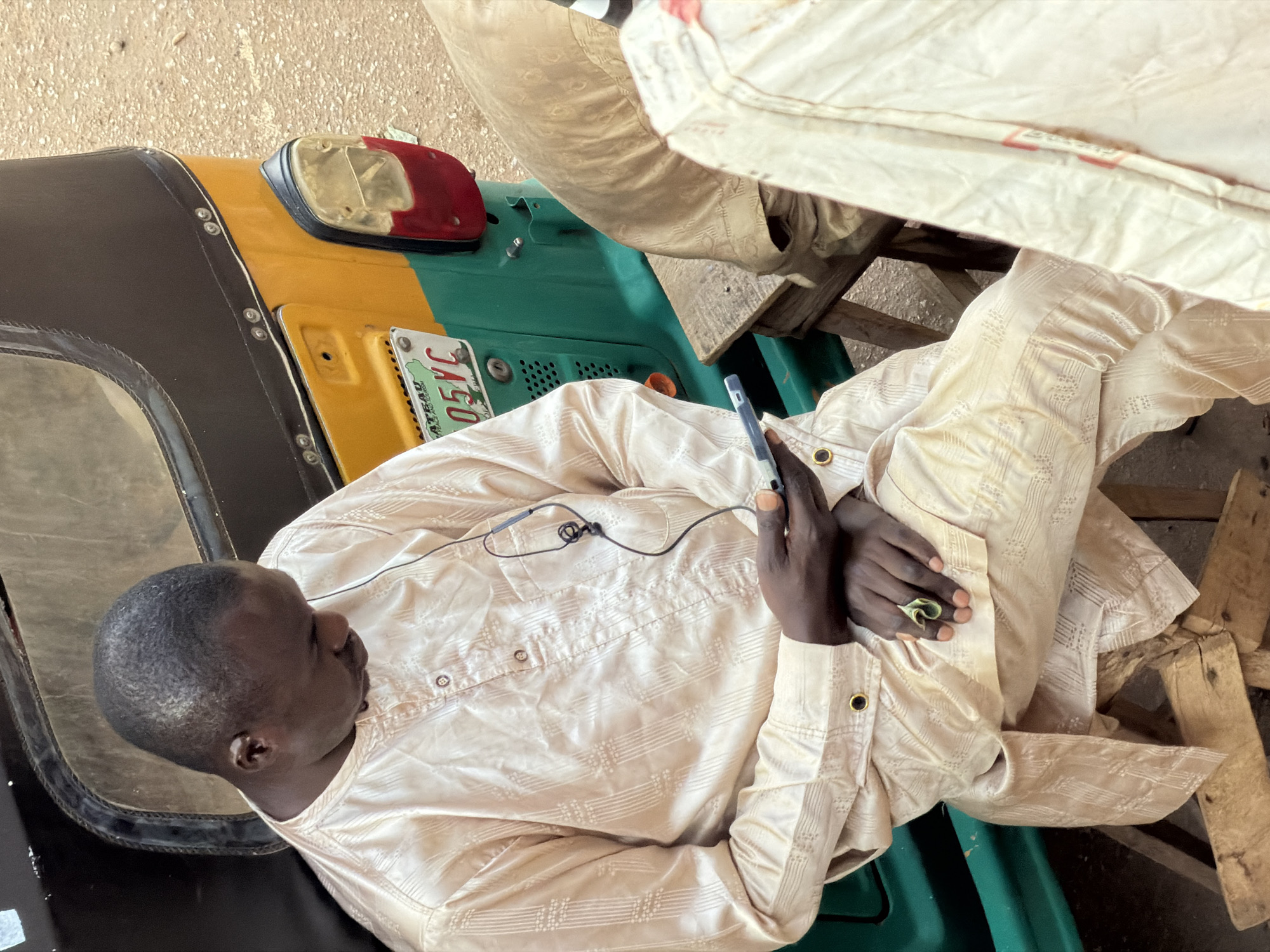 A man, wearing a light-colored traditional garment, sits on a wooden stool next to a yellow and green auto-rickshaw in Bukuru, Nigeria. He is looking down at a smartphone he holds in his hand, and appears to be listening to something through earphones. The man's left hand is resting on his lap, and a folded bill is visible between his thumb and forefinger. The auto-rickshaw has a license plate that reads "05VC." The ground is a rough, sandy texture. Another person, only partially visible in a similar garment, sits to the right of the man.