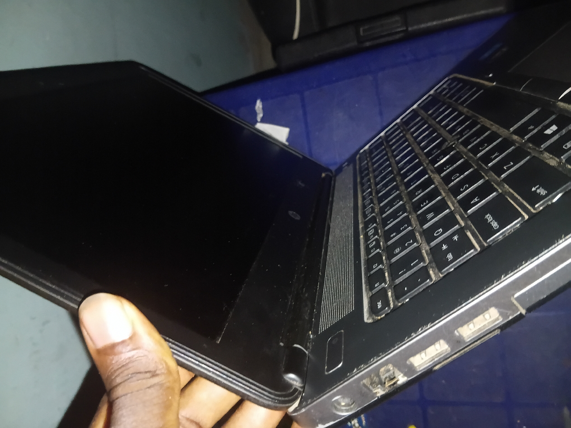 A person's hand is holding the edge of an open black laptop, partially obscuring the top left corner of the screen. The laptop is placed on a dark blue surface. The keyboard of the laptop is visible and appears to be dirty. The scene is indoors, and the lighting is dim. The location is Jalingo, Nigeria.