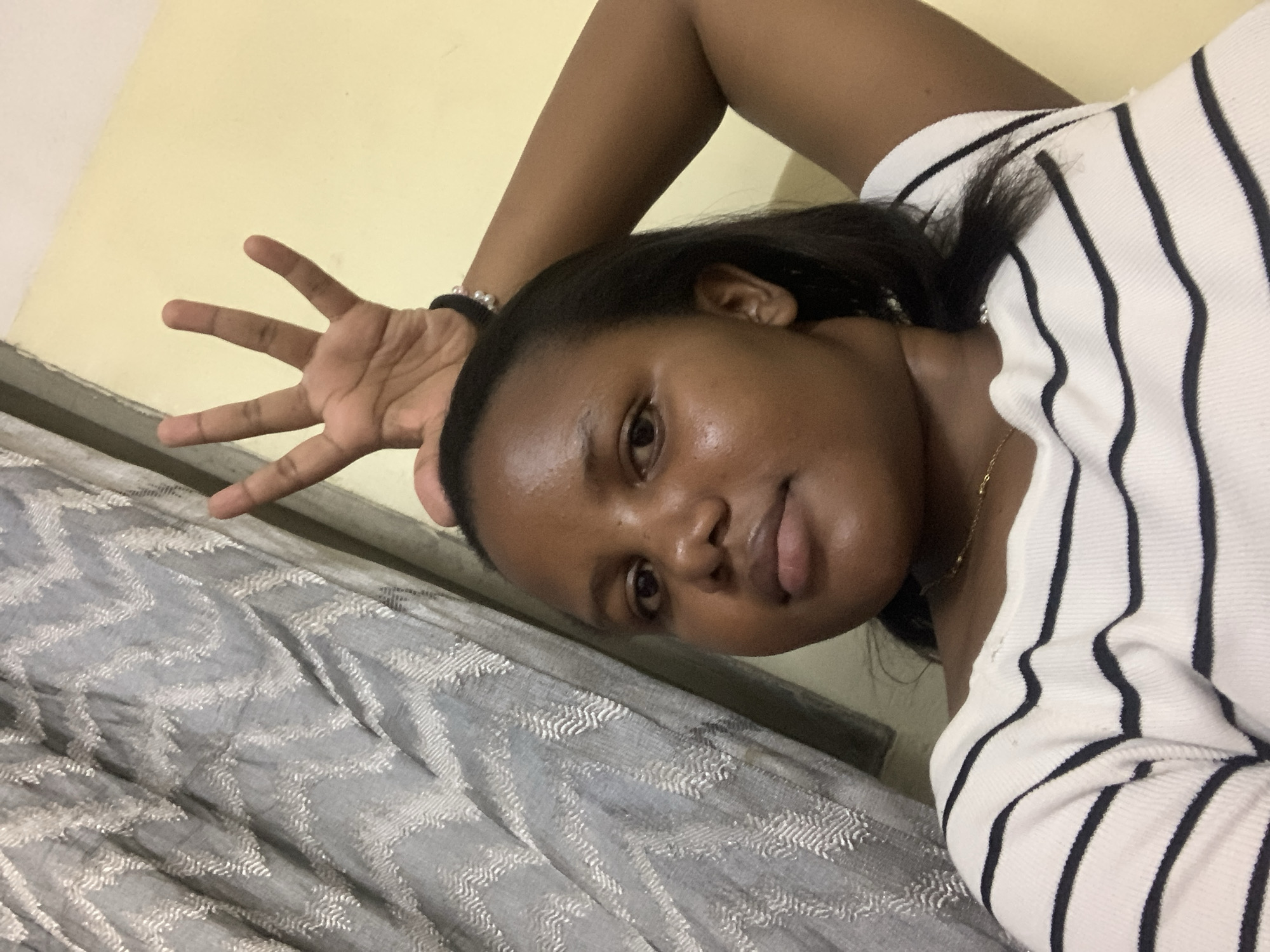 A young woman with dark hair and skin is lying down on a bed with a patterned grey blanket. She is wearing a white top with black stripes. Her right hand is raised with her fingers splayed. She is indoors, in a room with a yellow wall. The image provides no information to determine the location in Dar es-Salaam, Tanzania.